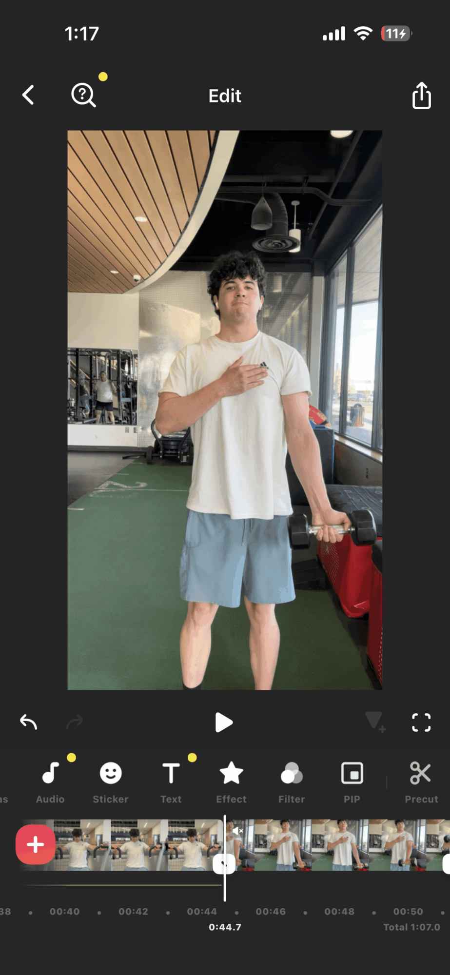Young man standing indoors, wearing a white t-shirt and blue shorts, holding a phone, with large windows behind him.