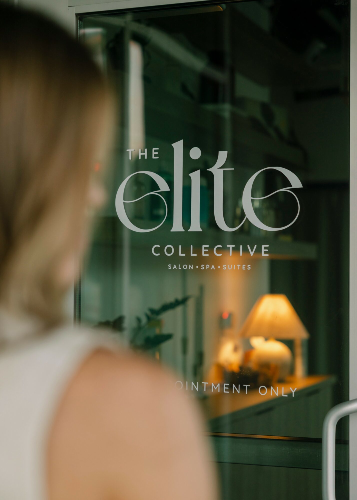 Blurred person looking at glass door with logo for 'The Elite Collective' salon and spa, interior with lamp visible inside.