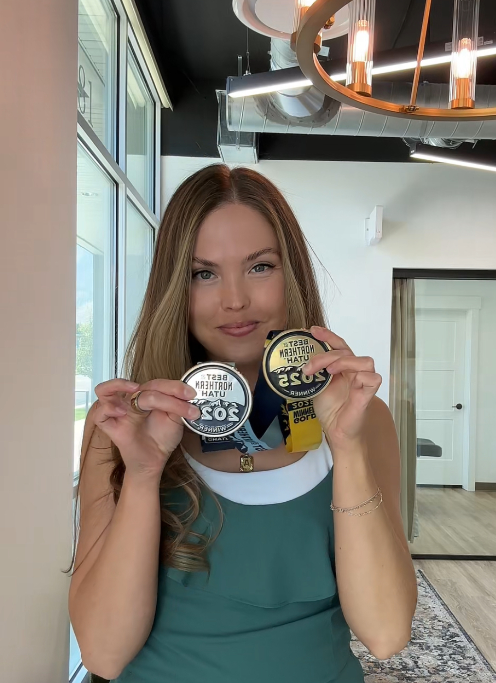 Woman holding two medals, smiling indoors with modern decor and large windows behind her.