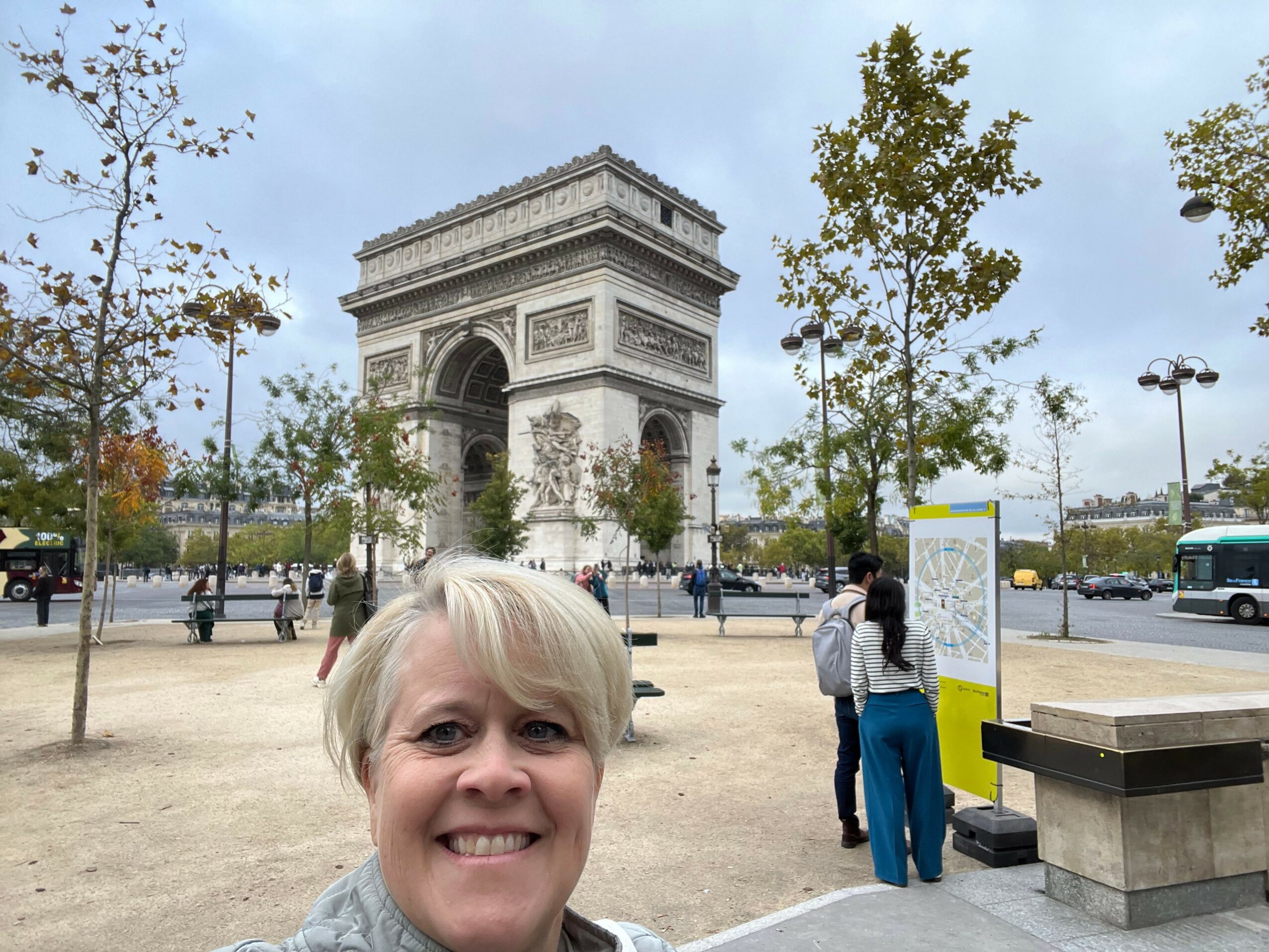 A smiling woman with glasses and blonde hair stands outdoors near the Arc de Triomphe in Paris, with trees and a cloudy sky.