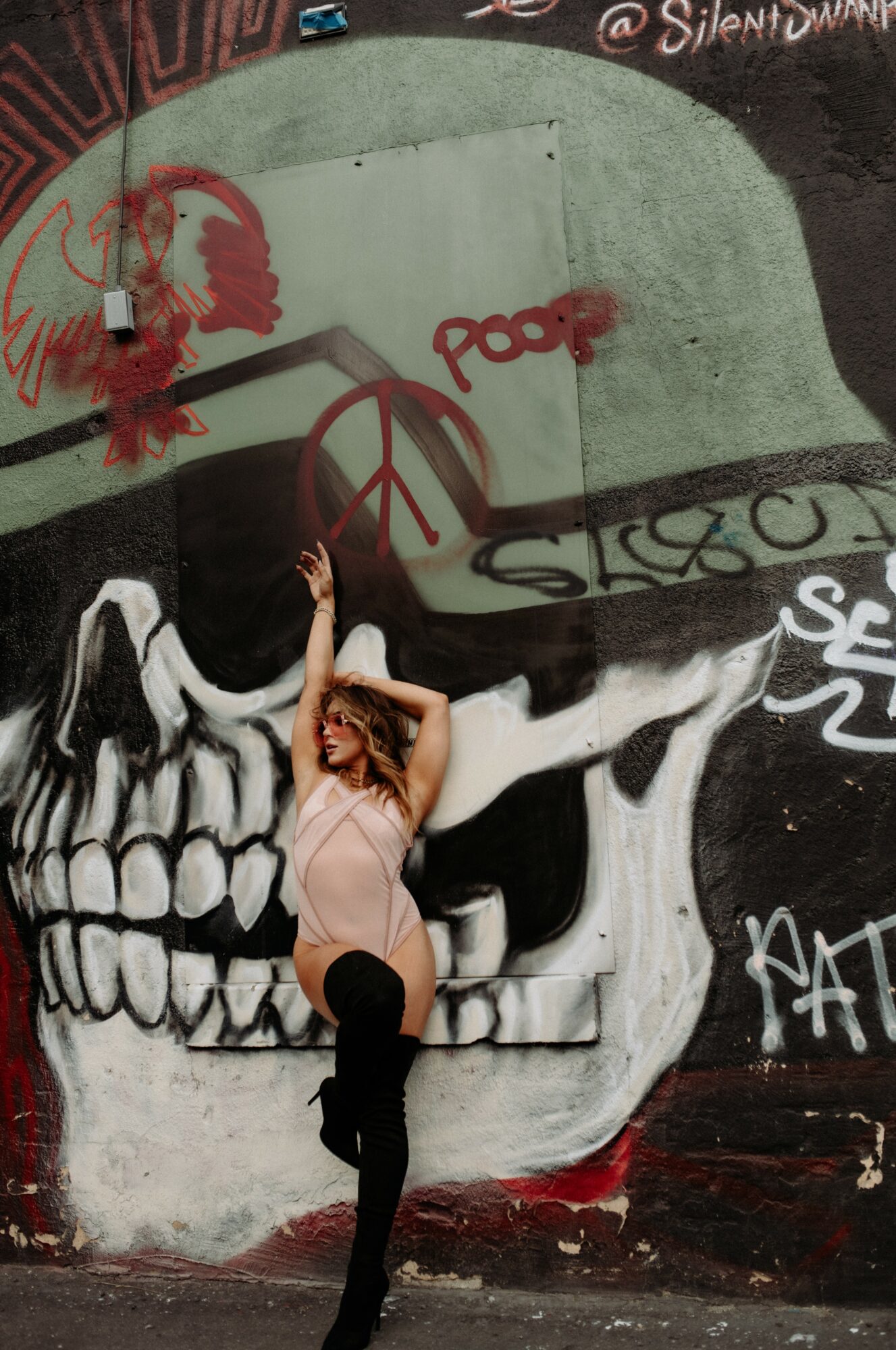 Person with curly hair poses in front of a large skull graffiti mural with peace symbols and red spray paint, arms outstretched.
