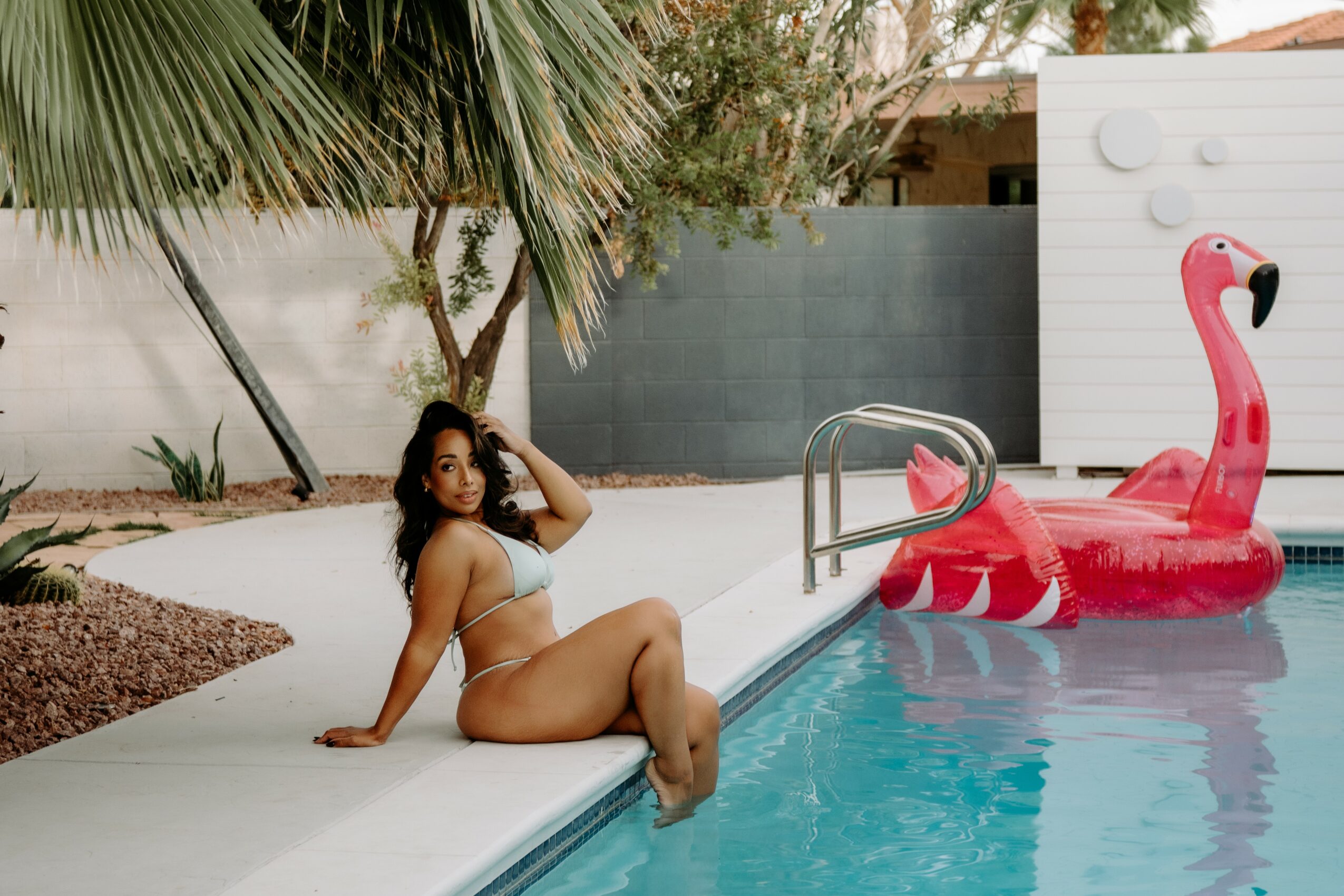 Woman in a bikini sitting on pool edge next to a pink flamingo float, with trees and a wall in background.