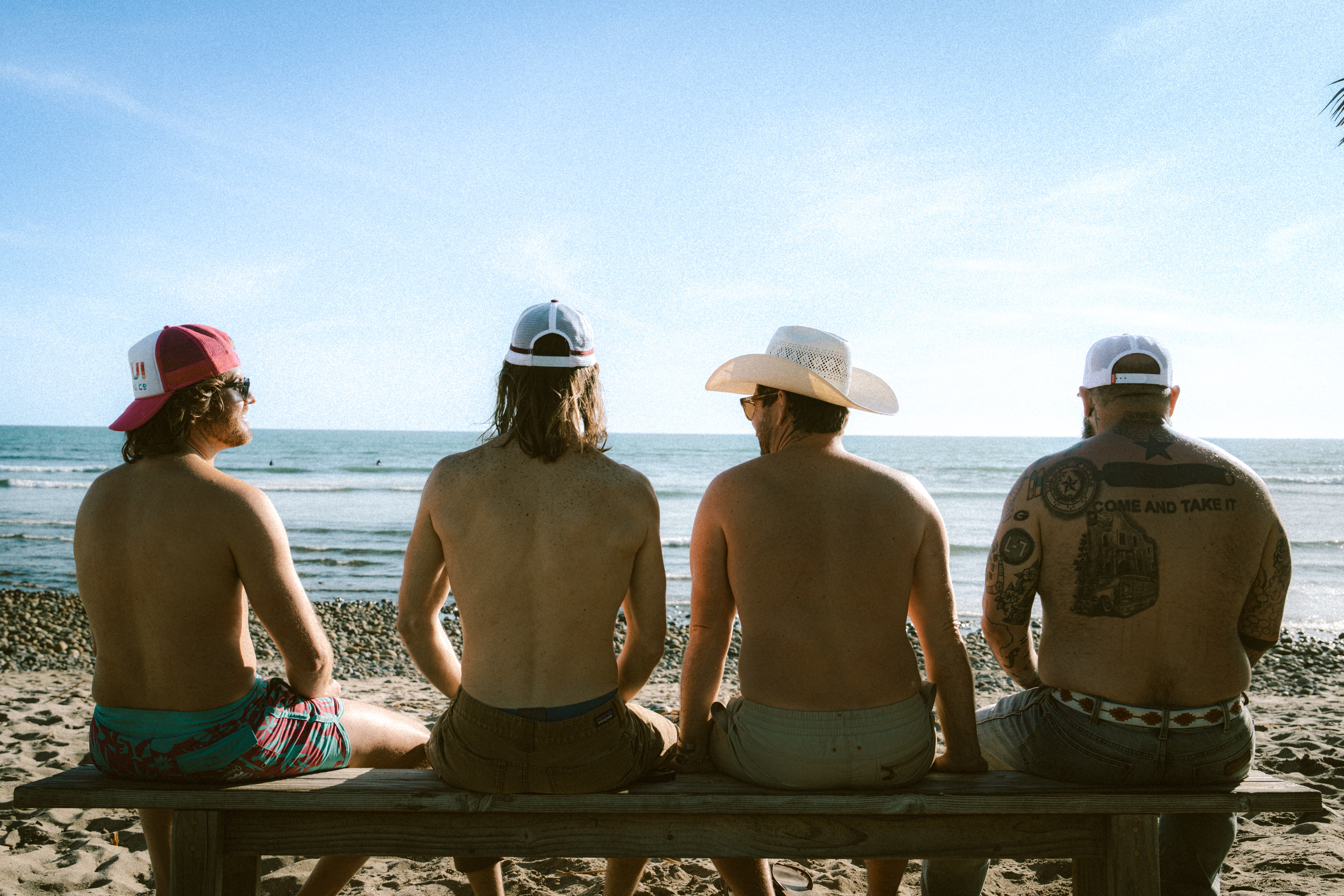 Four people sit on a bench facing the ocean on a sunny beach, wearing hats and swimwear.
