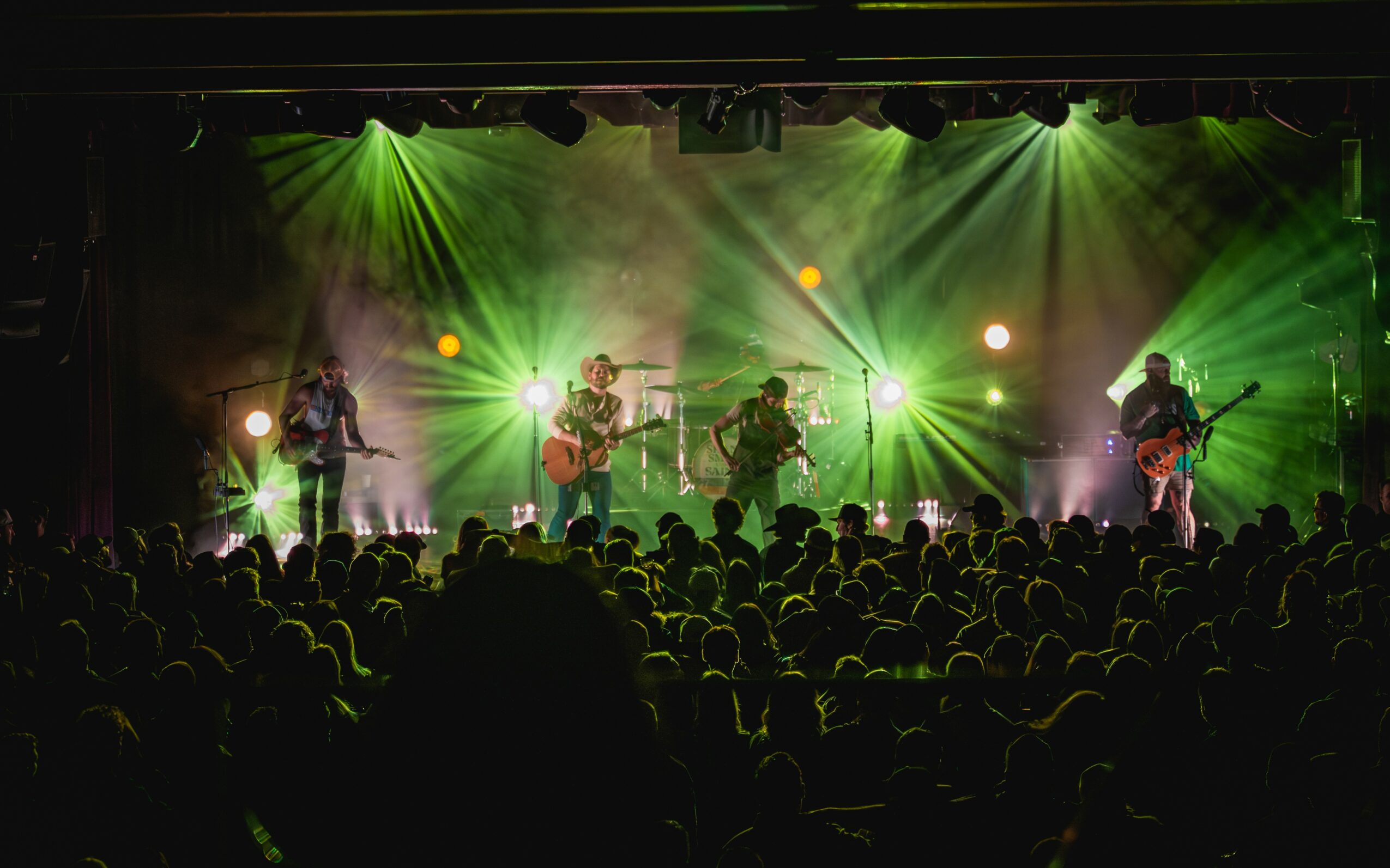 Band performs on stage with green and yellow lighting, audience watching attentively, in a concert setting.