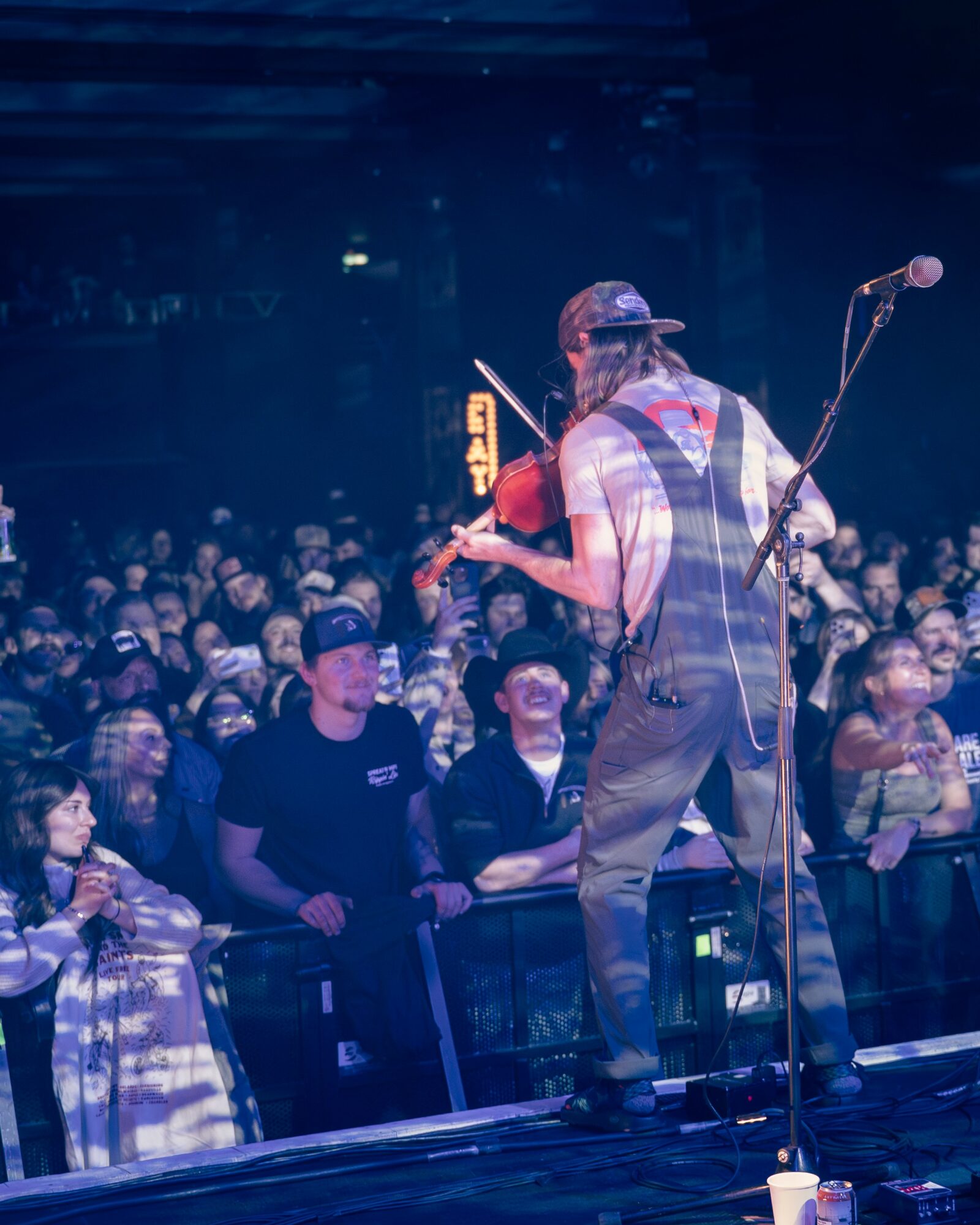 Musician playing violin on stage with audience watching, microphone stand nearby, dark background, stage lighting.