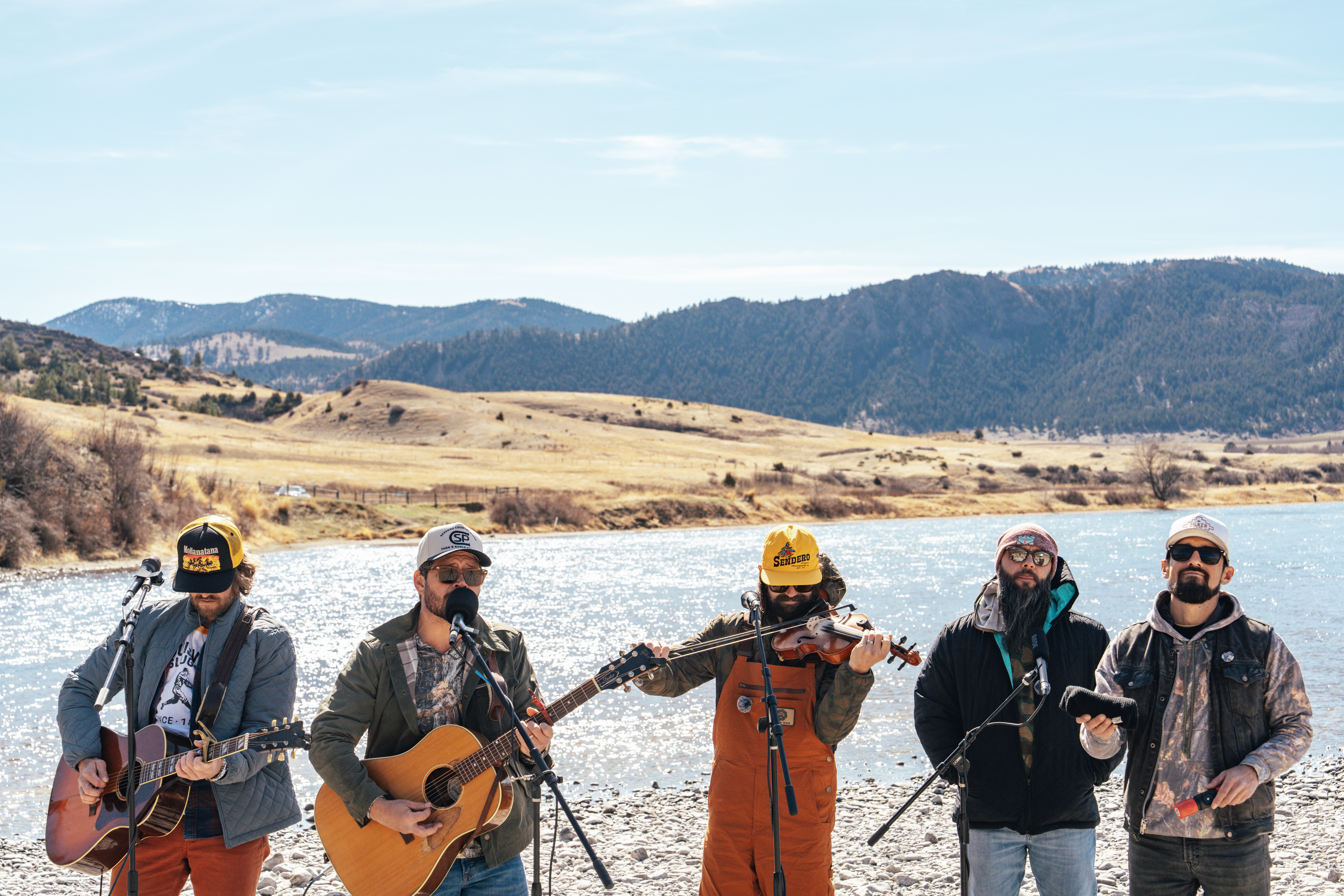 Four people playing guitars outdoors near a river with mountains in the background.