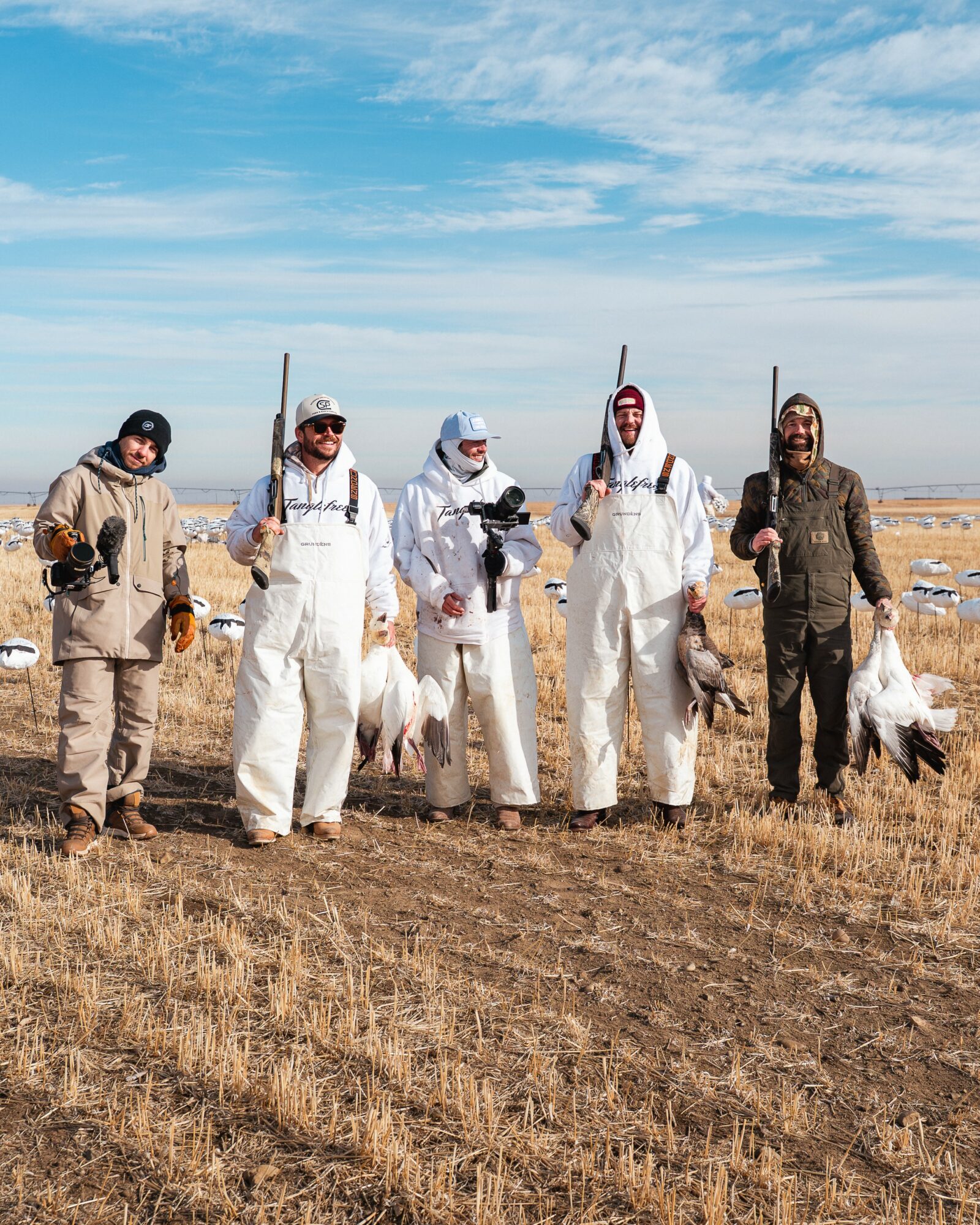 Five people stand outdoors in a field holding hunting rifles and birds, with a blue sky and clouds above.