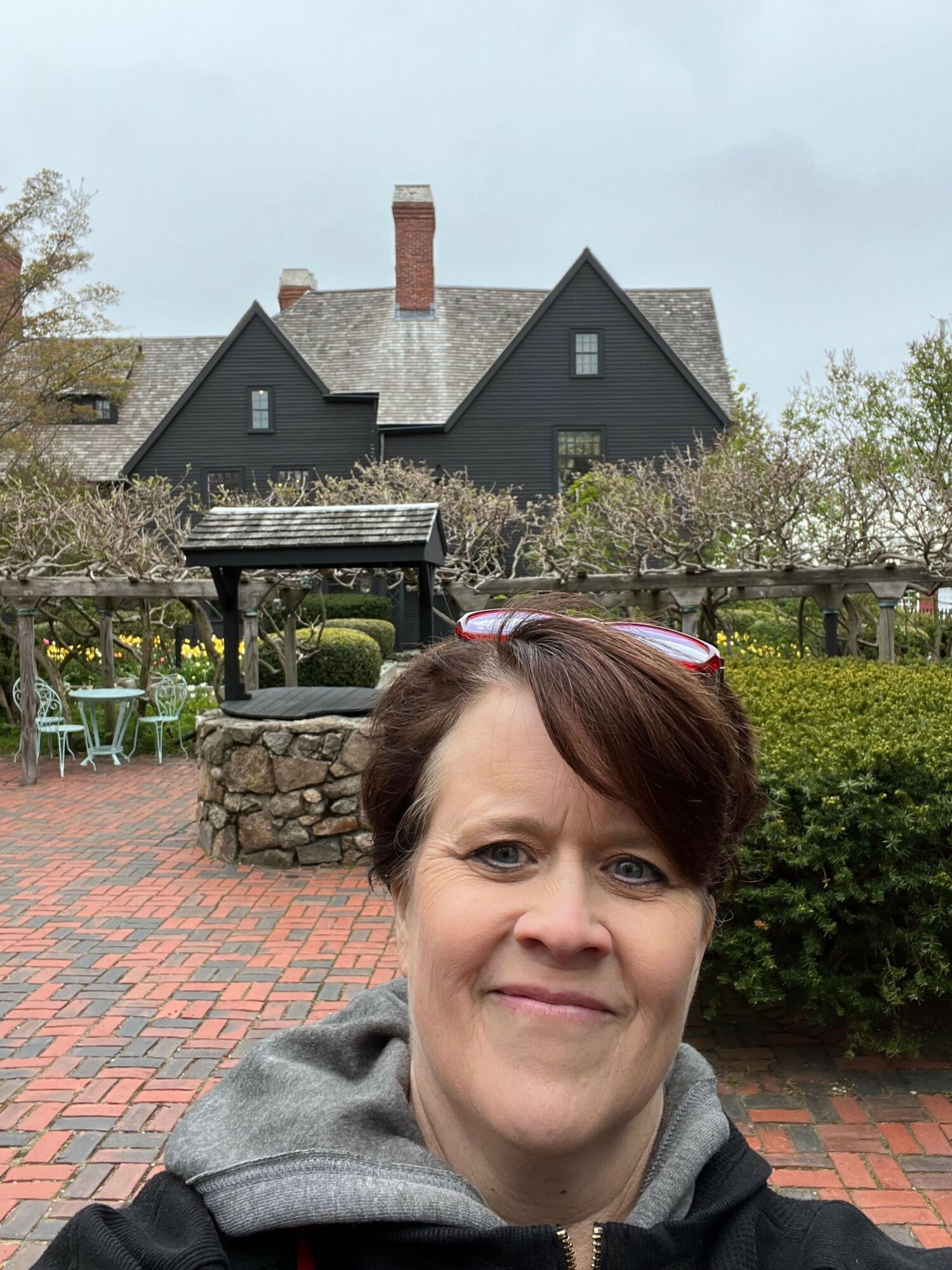 Woman with short brown hair smiling outdoors on brick pathway, house with chimney and trees in background.