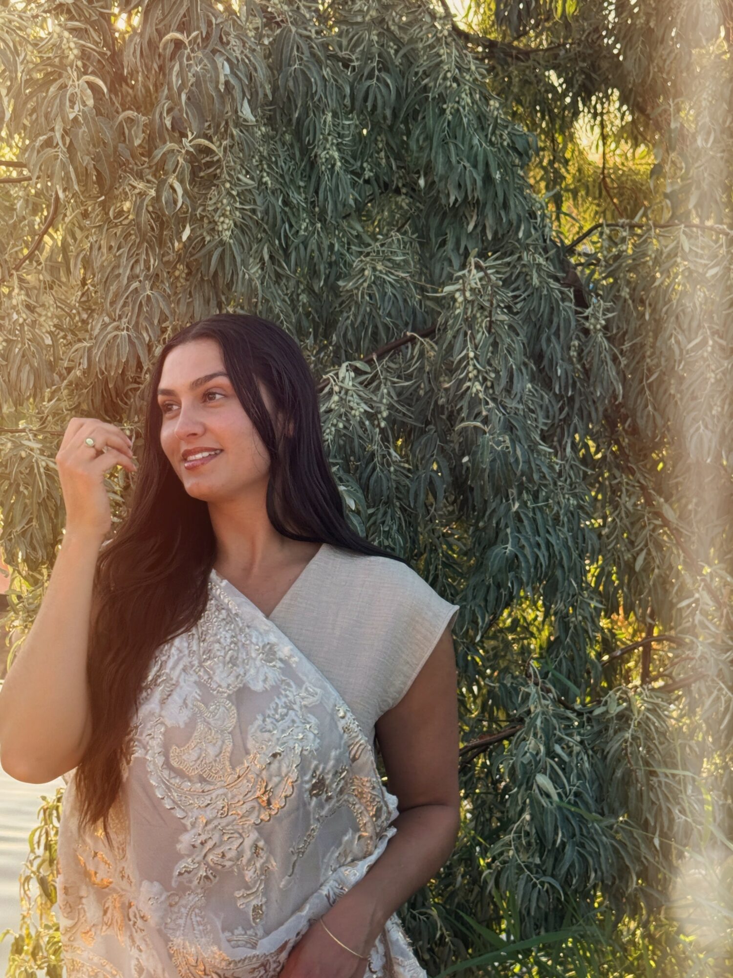 Woman with long dark hair smiling outdoors, standing in front of green trees, sunlight shining through leaves.