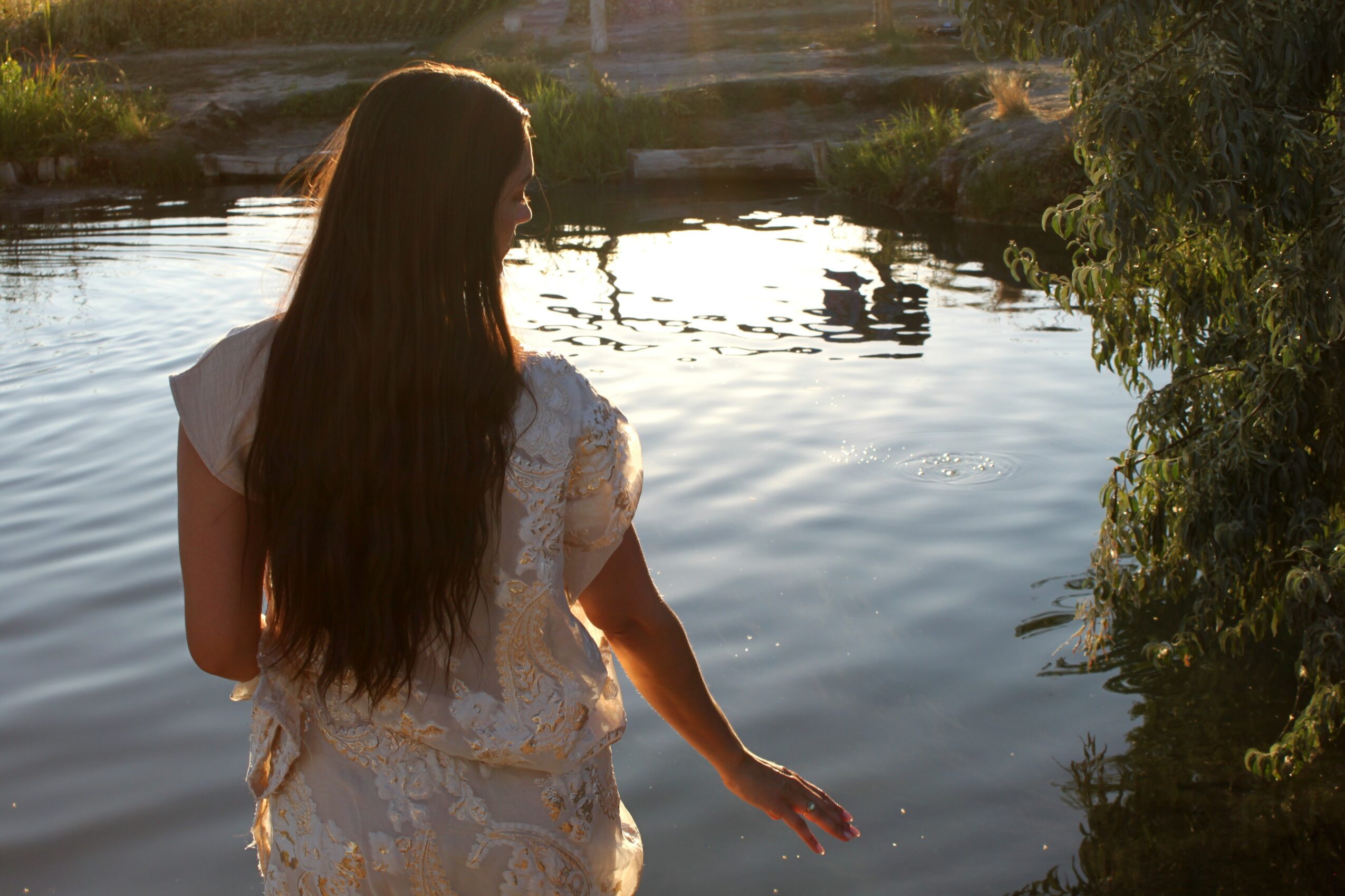 Woman with long hair standing by a body of water, touching the surface with her hand, surrounded by trees and sunlight.