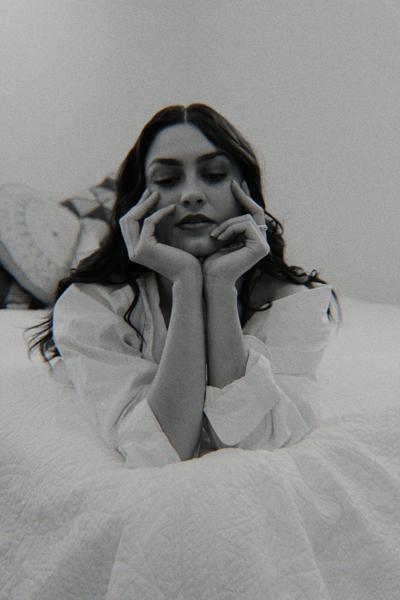 A woman with long dark hair rests her chin on her hands, looking down thoughtfully, in a monochrome setting.