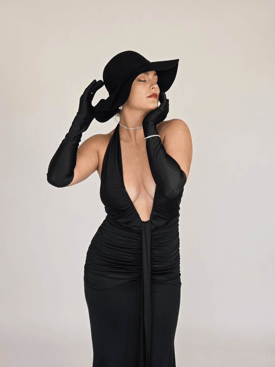 Woman in black dress and large hat, posing with eyes closed, against plain background.
