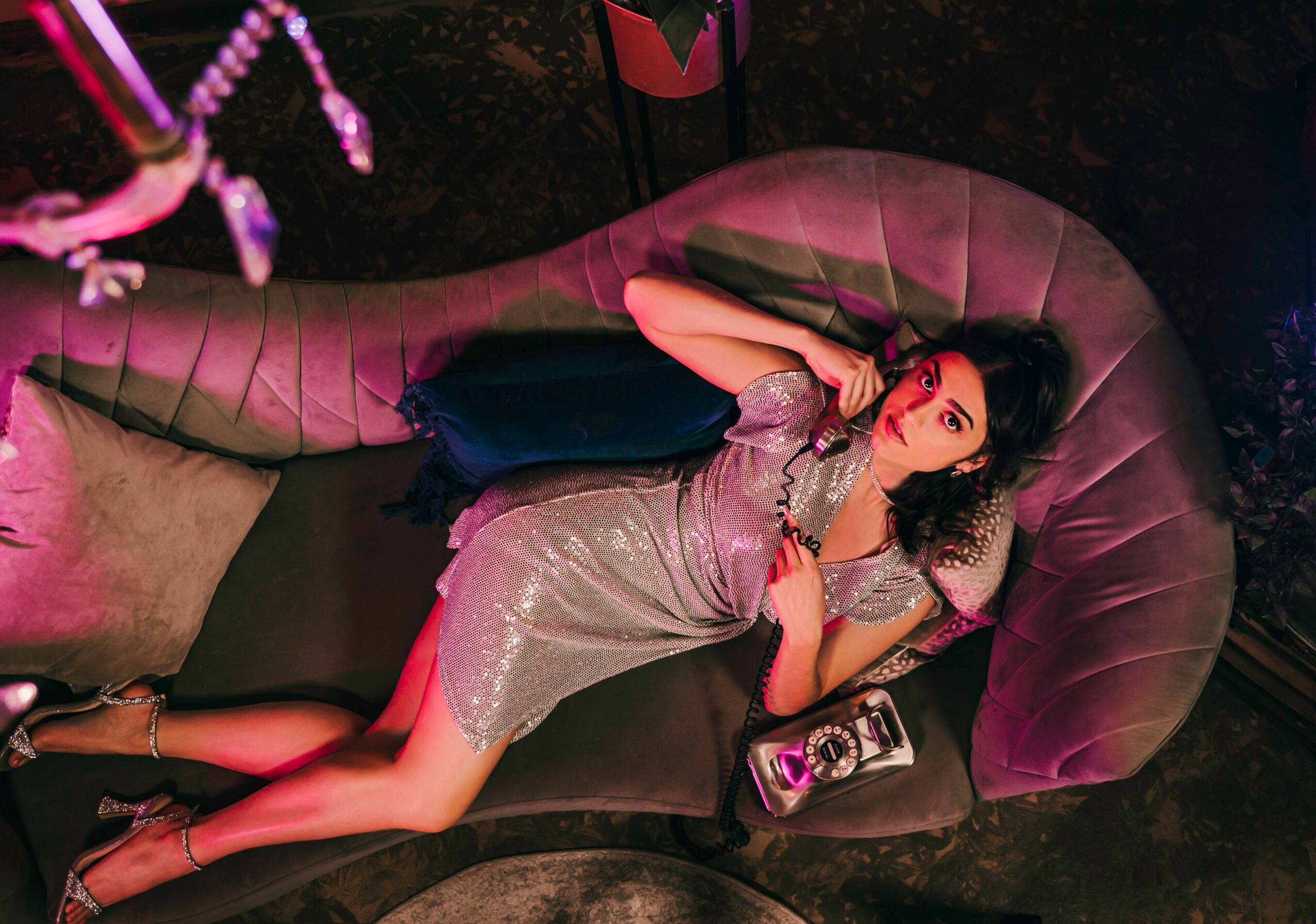 Woman lying on a curved sofa, wearing a silver dress, holding a phone, with a dark background and pink lighting.