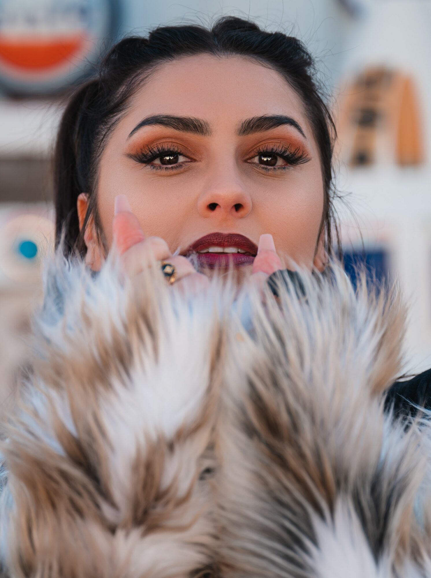 Close-up of a woman with makeup, holding a furry object partially covering her face, in an indoor setting.
