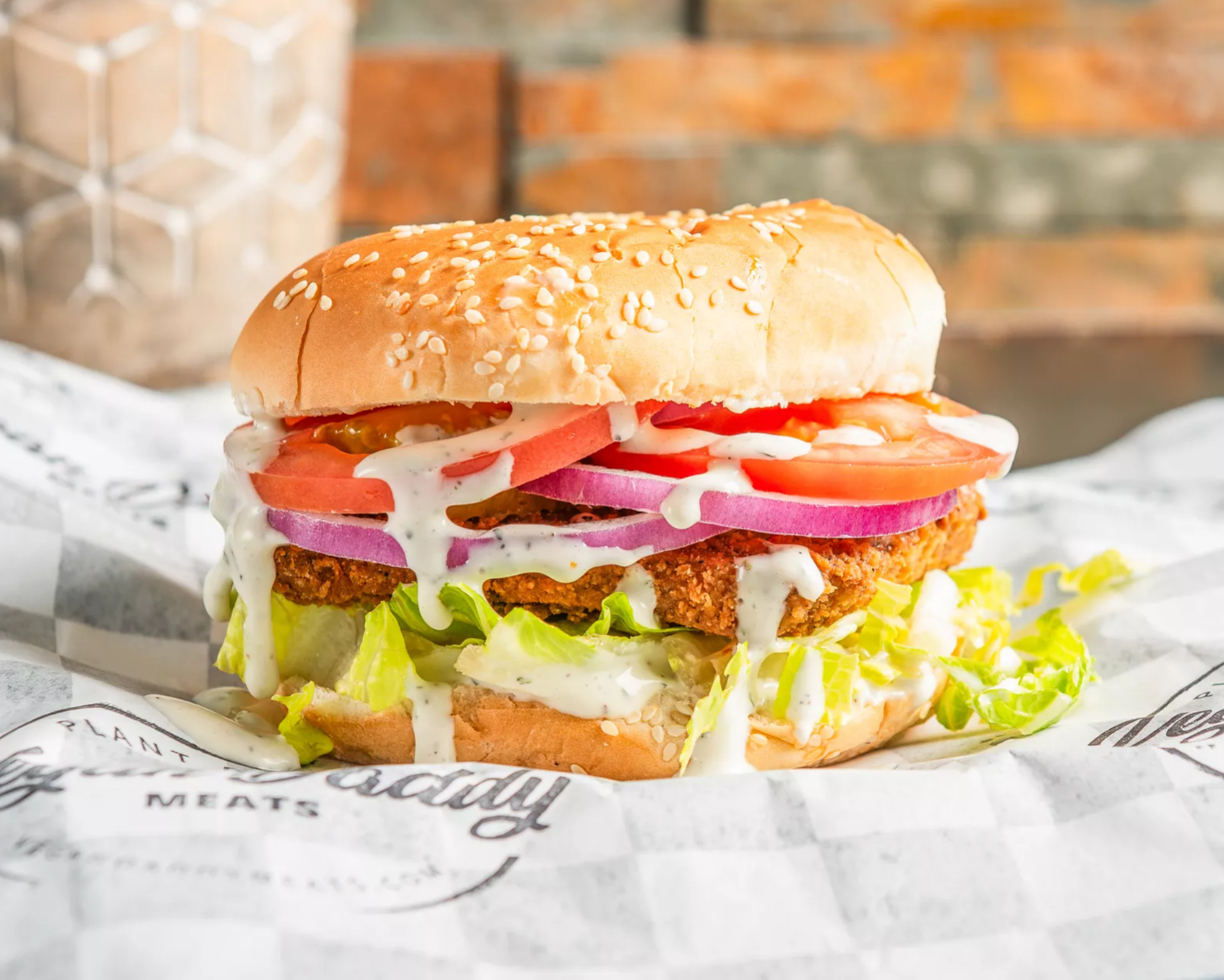 Sandwich with lettuce, tomato, onions, pickles, and sauce in a sesame bun on paper, brick wall background.