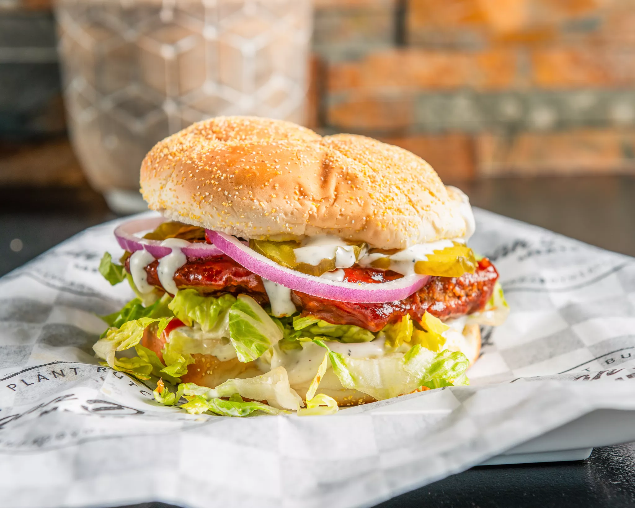 Sandwich with lettuce, pickles, onions, tomato, and condiments in a bun on paper, brick wall background.