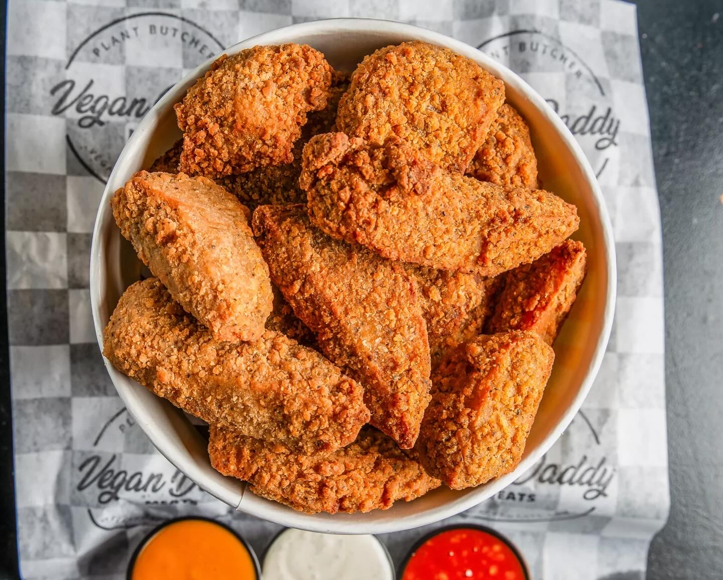 Bowl of fried chicken pieces on checkered paper with dipping sauces below, viewed from above.