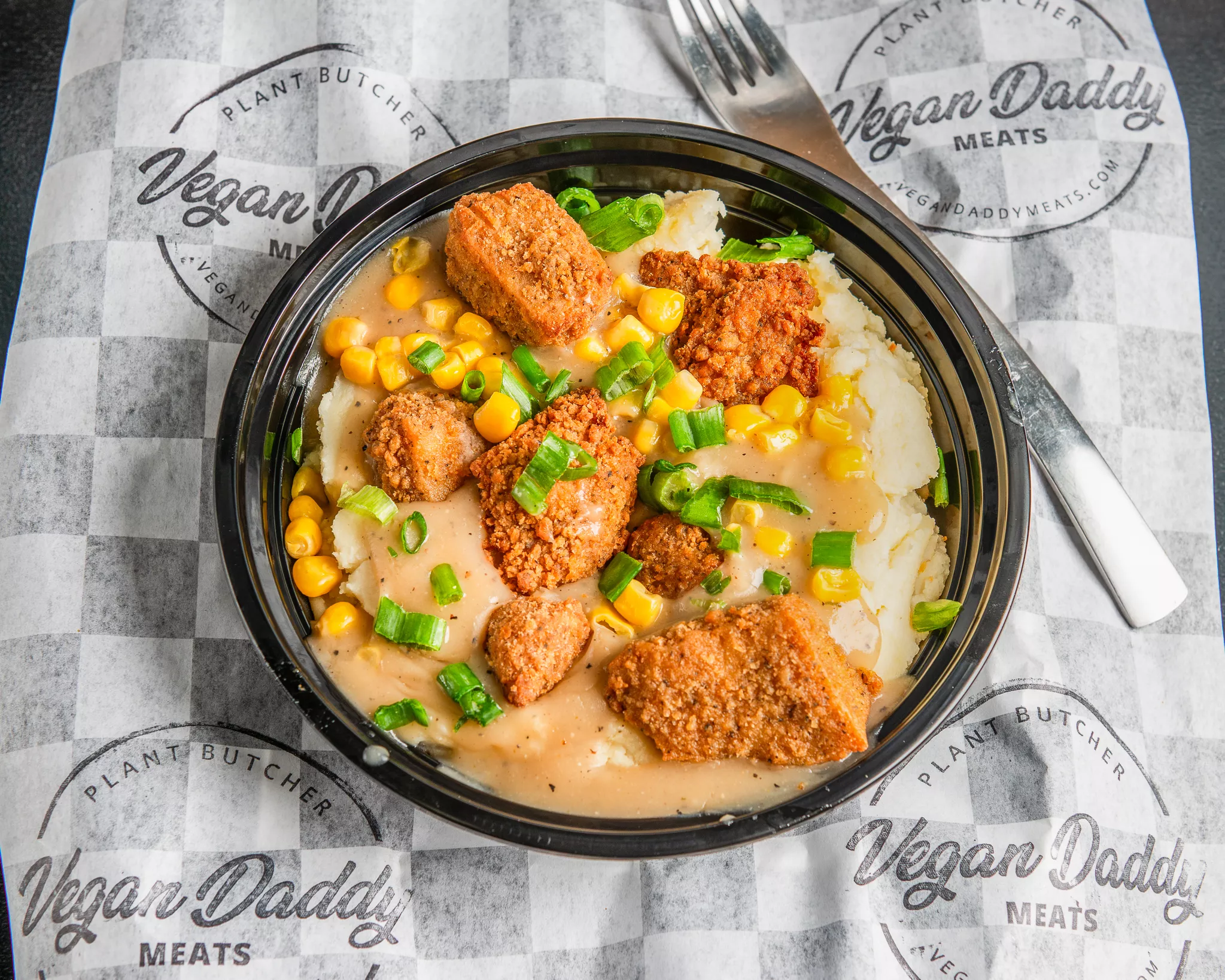 Bowl of fried plant-based chicken pieces with rice, corn, and green onions, on branded paper with a fork nearby.