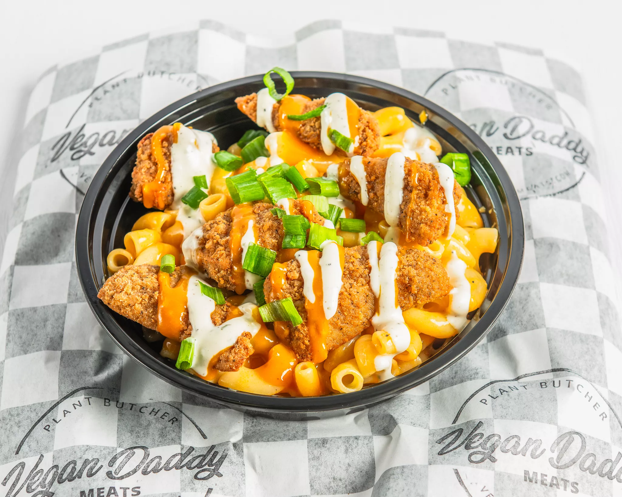 Bowl of macaroni and cheese topped with fried chicken pieces and chopped green onions, served on checkered paper.