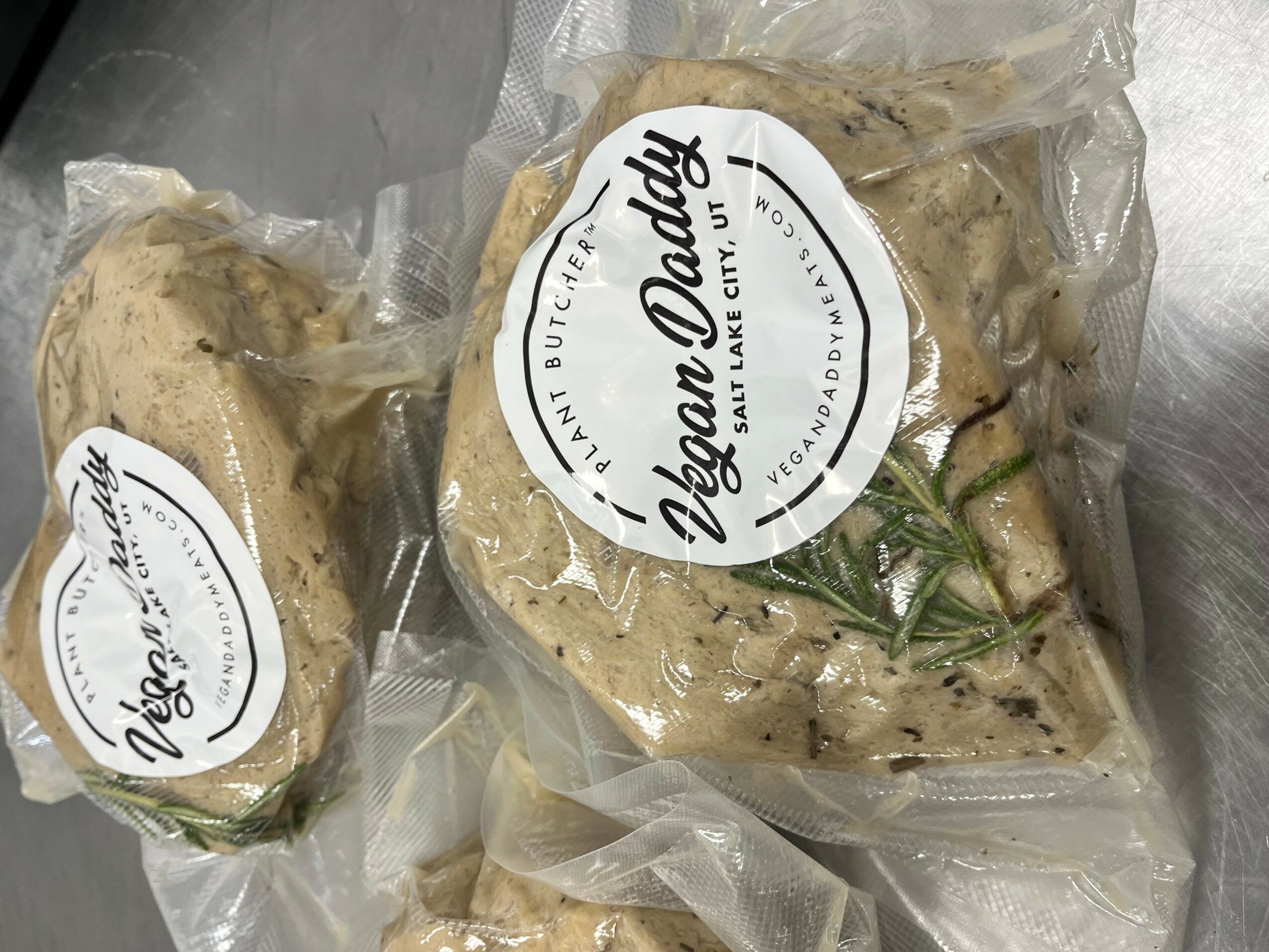 Two vacuum-sealed packages of vegan chicken, labeled Zoe Daddy, with herbs inside, on a metallic surface.