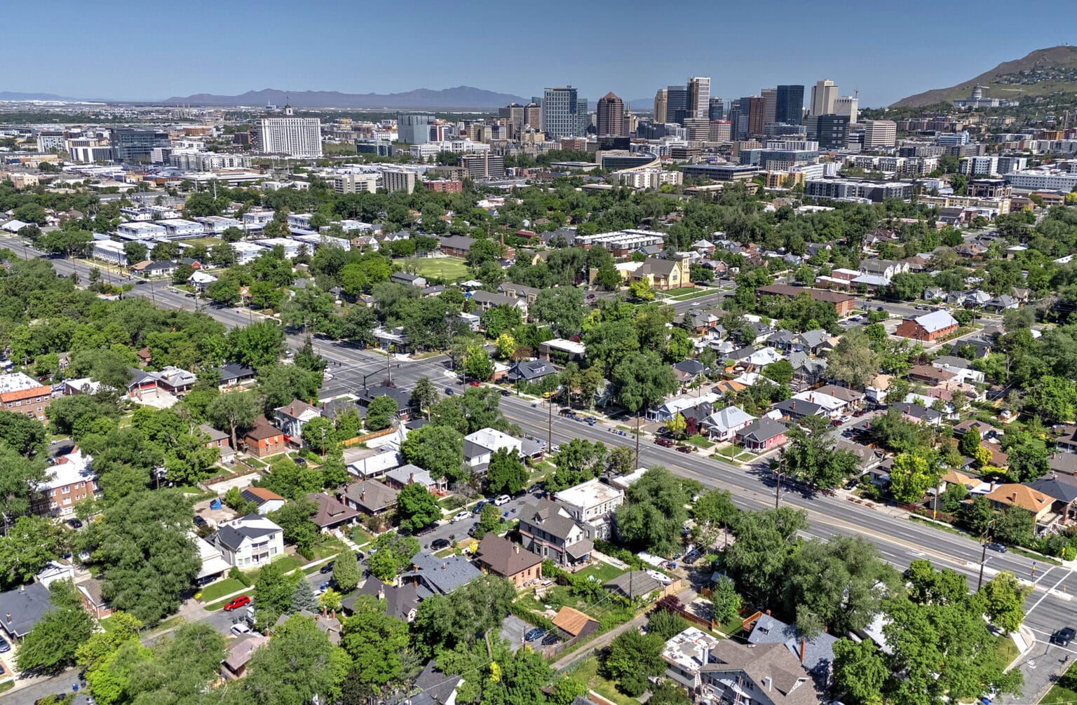 Aerial view of a city with a mix of residential houses, trees, and a downtown skyline in the background under a clear blue sky.