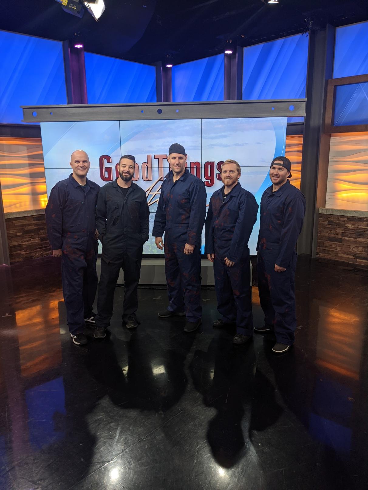 Five men stand together in a studio with a large screen behind them displaying text and graphics.