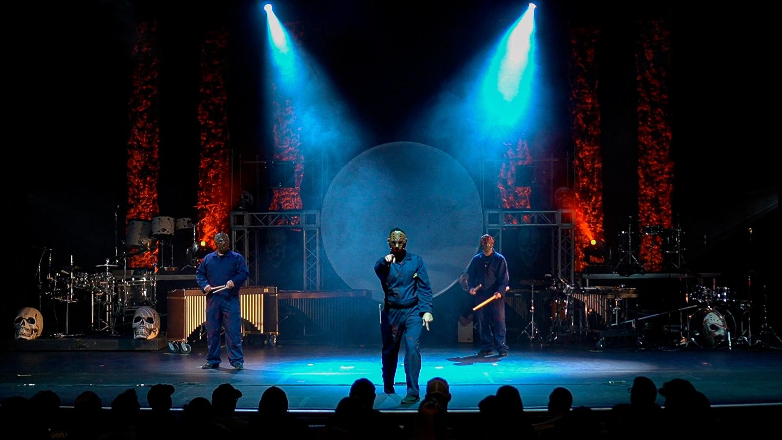 Three performers on stage with blue and red lighting, musical instruments, and skull decorations in the background.