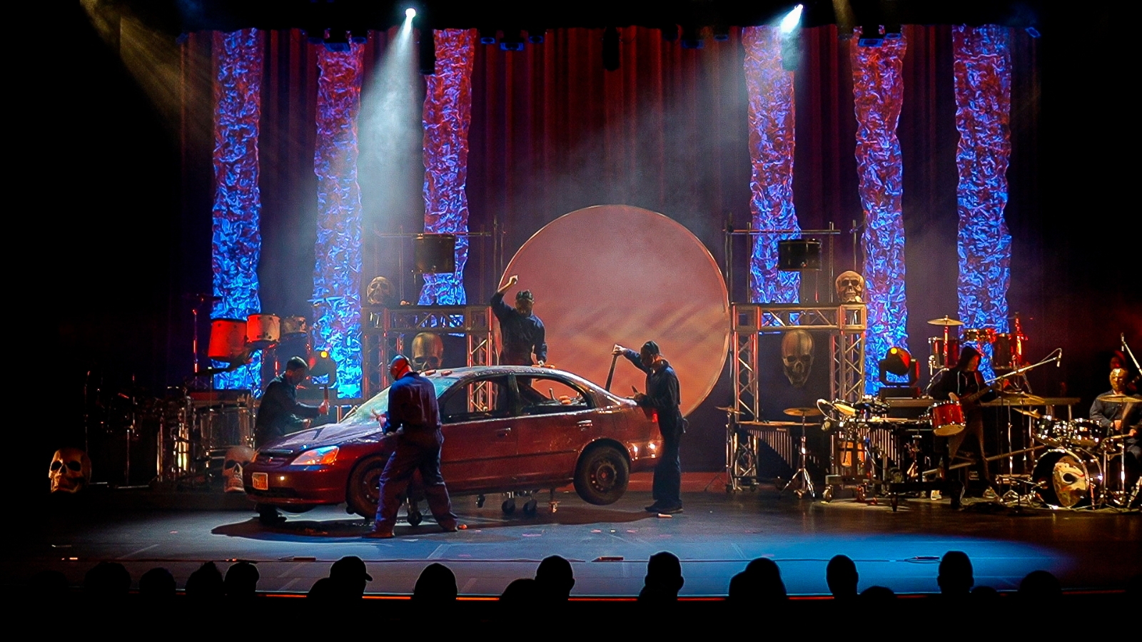 Stage with performers, a car, colorful lights, and a large circular backdrop, with audience silhouettes at the bottom.