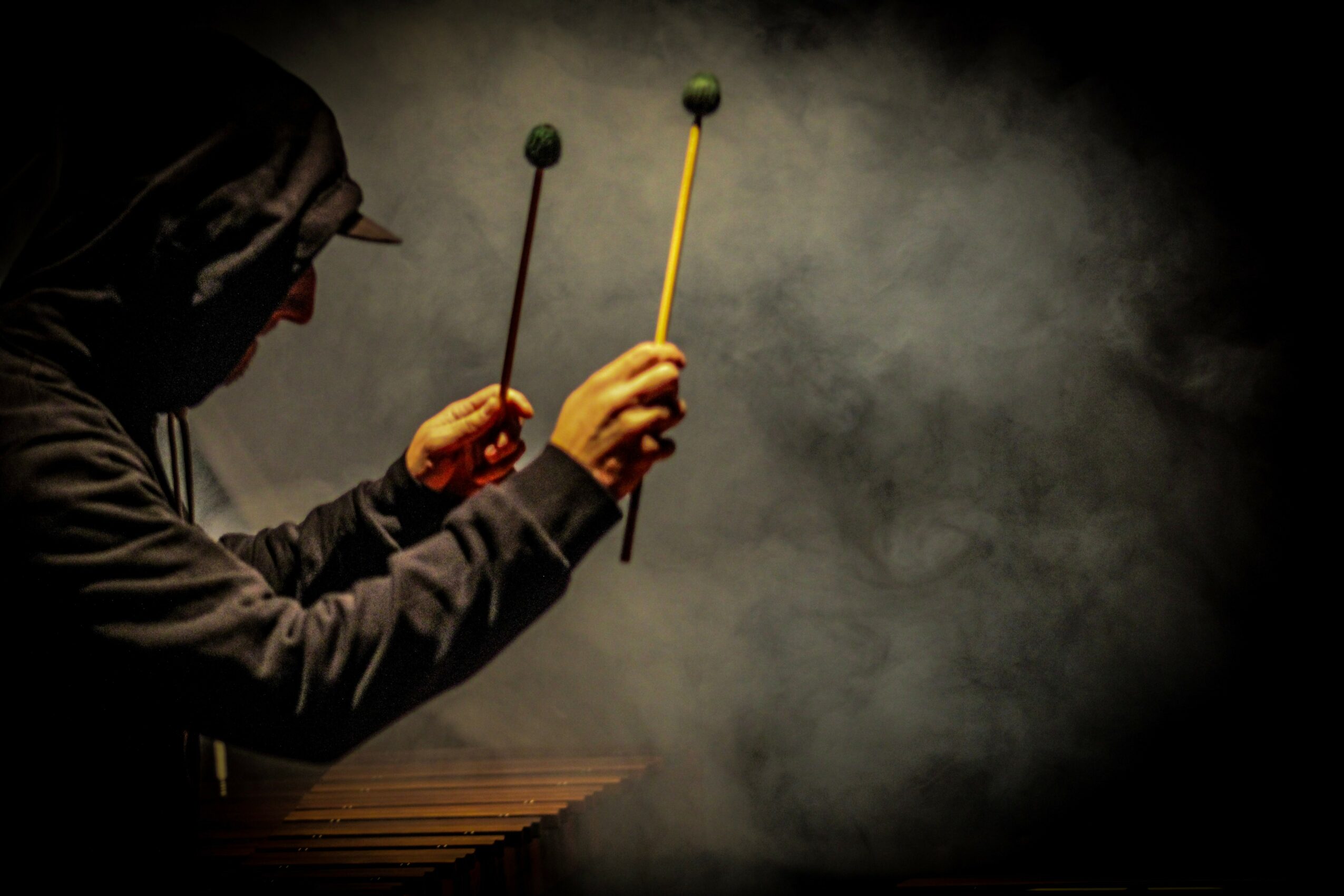 Person wearing a hoodie playing drums with mallets, surrounded by dark smoke or fog, in a dimly lit setting.