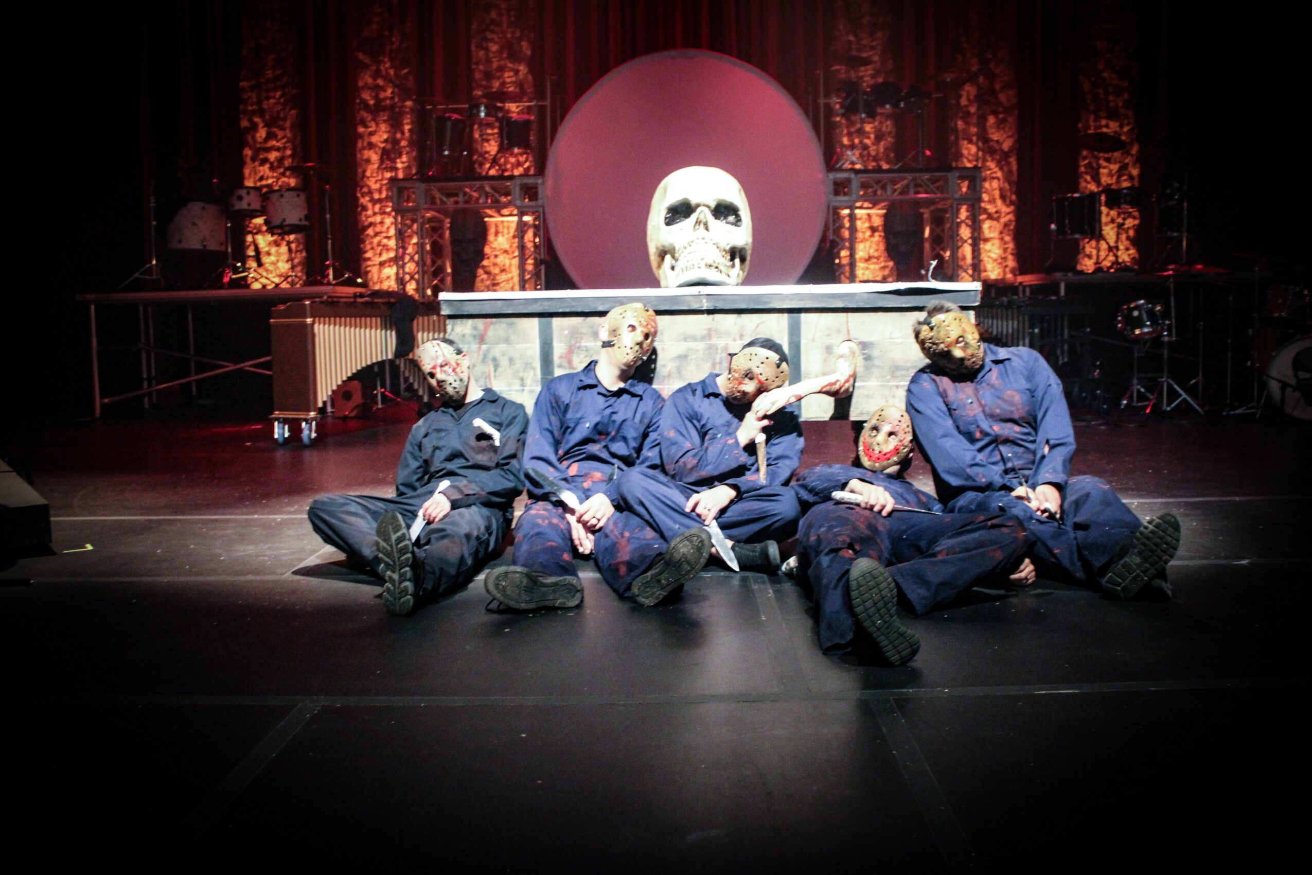 Four people sit on the stage floor in front of a large skull and circular backdrop, with a dark curtain behind them.