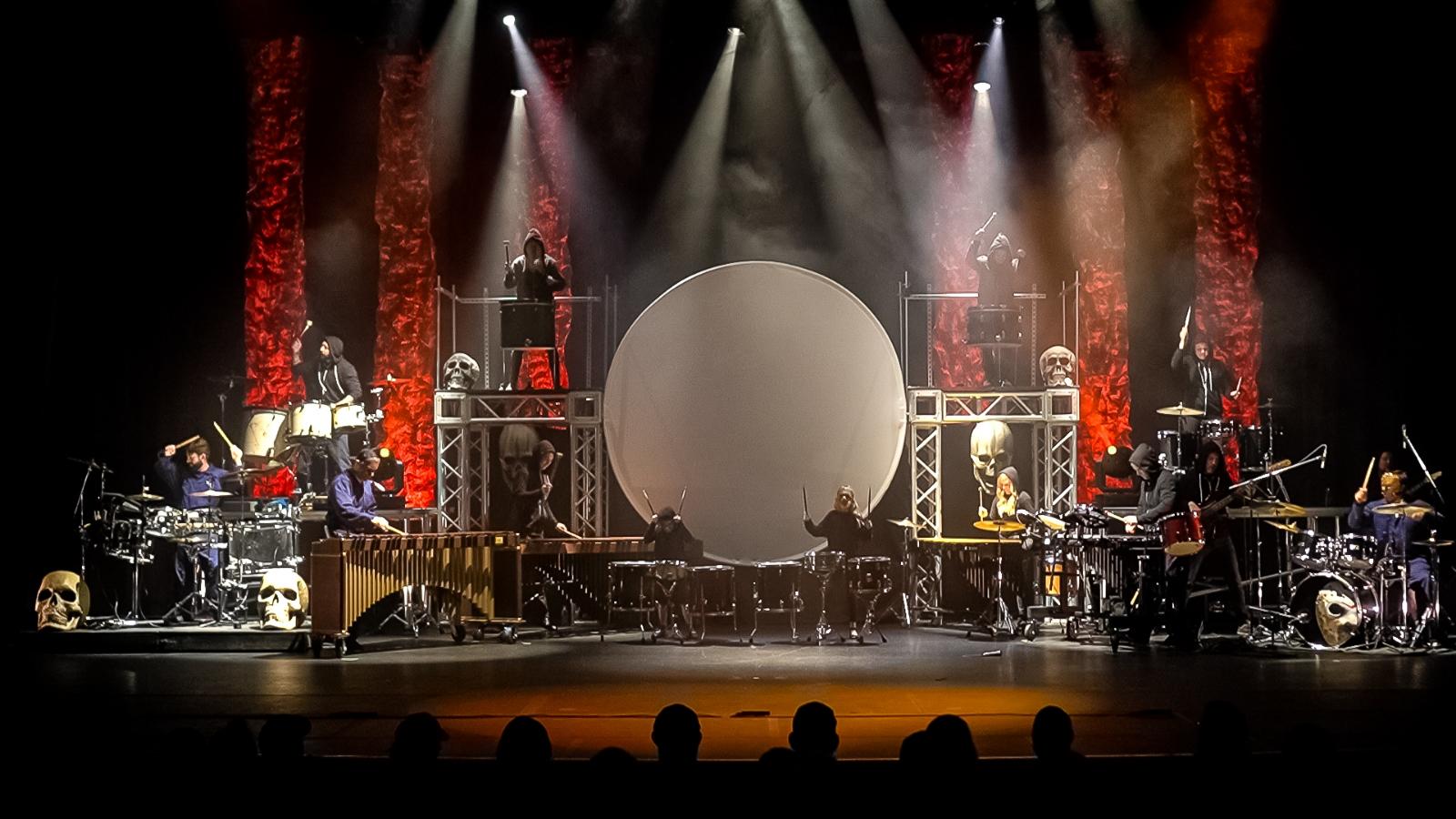 Musicians perform on stage with drums, keyboards, and a large circular screen, illuminated by spotlights and red vertical light columns.