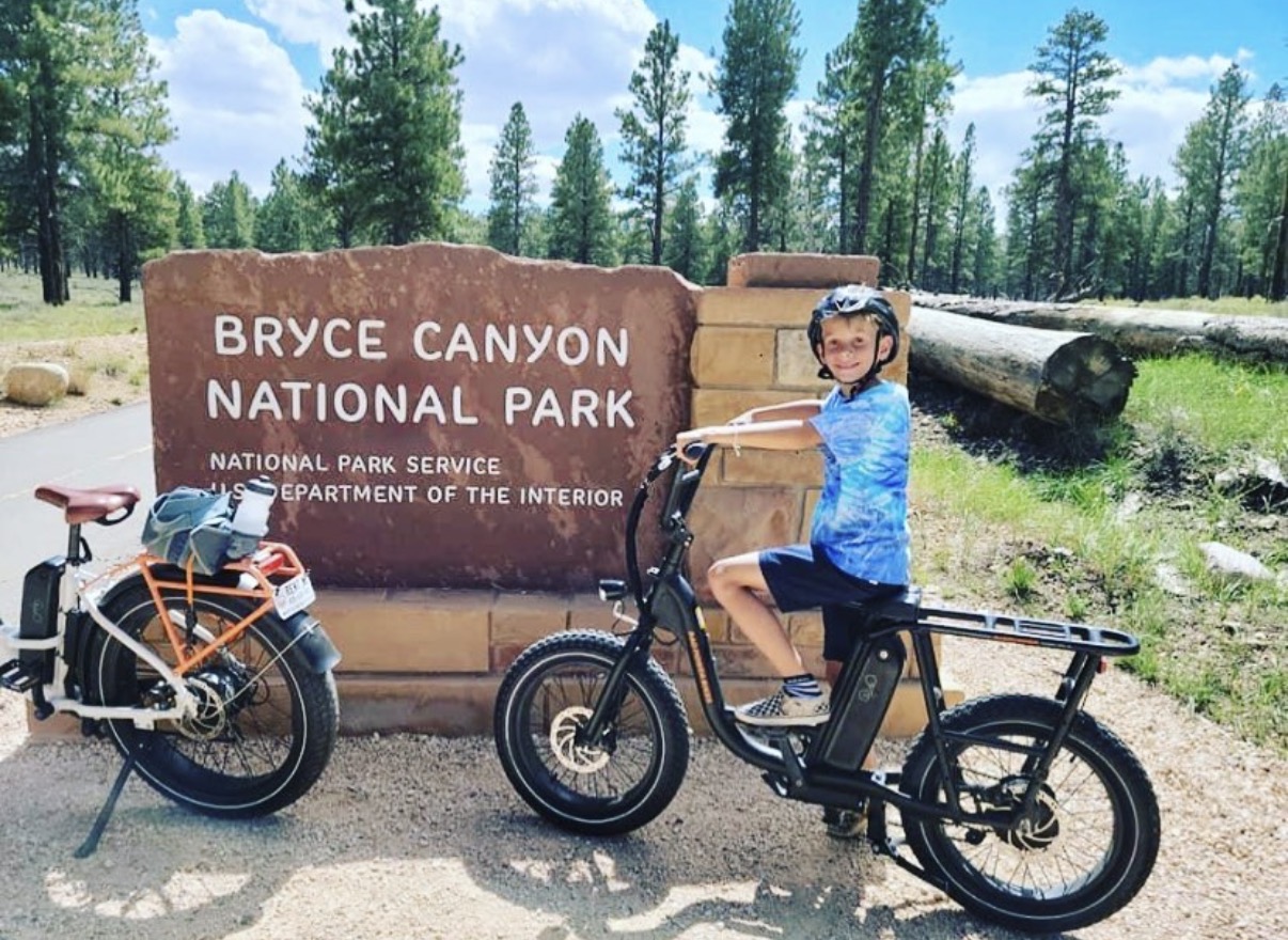 Child wearing a helmet sitting on an electric bike at Bryce Canyon National Park entrance sign, with a second bike nearby, trees in background.