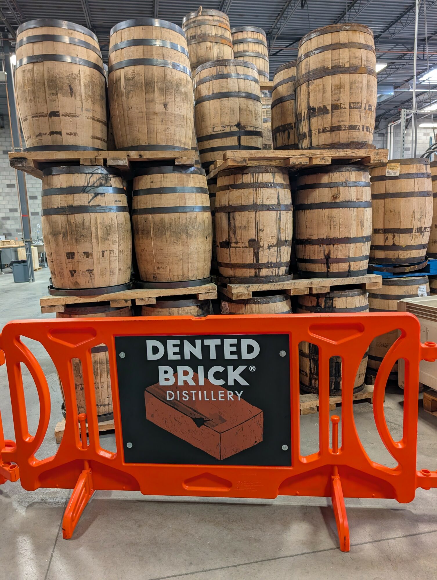 Stacked wooden barrels behind a black sign with white text and an orange barrier in front, in a distillery setting.