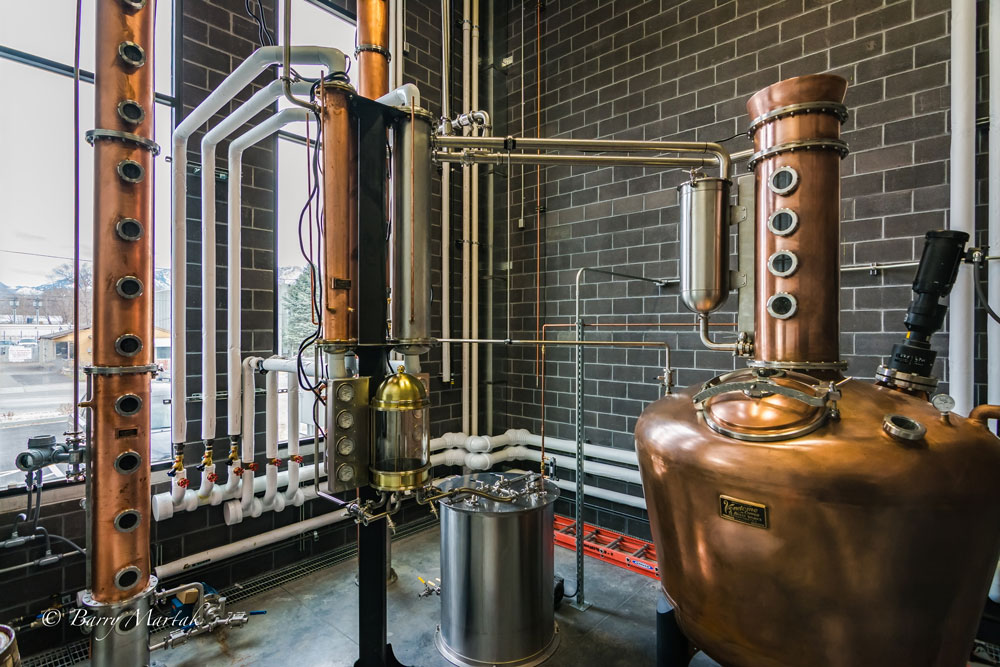 Industrial distillation apparatus with copper and glass components against a brick wall, pipes, and gauges, in a laboratory setting.