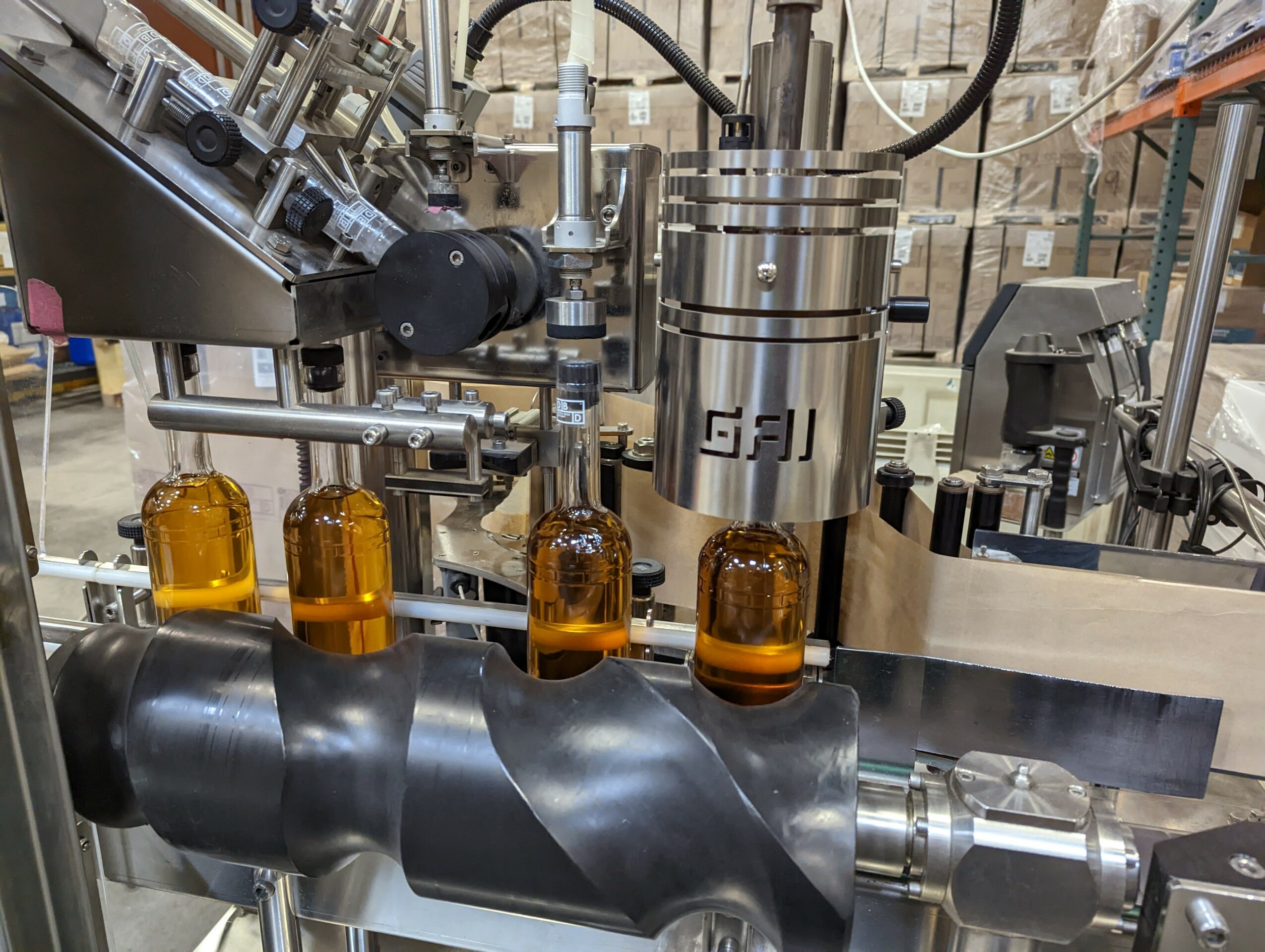 Industrial machinery filling amber bottles with liquid, with various pipes and mechanical components visible.