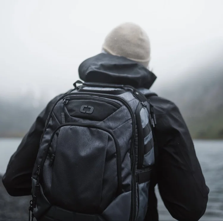 Person wearing a beige beanie and black jacket with a large black backpack, facing away outdoors on a cloudy day.