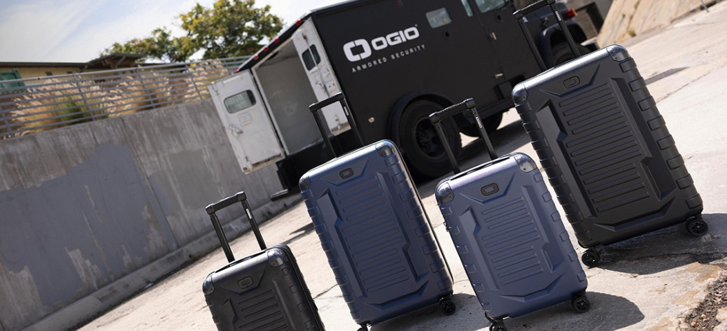 Four suitcases of different sizes and colors are placed outdoors near a black vehicle, with a concrete wall in the background.