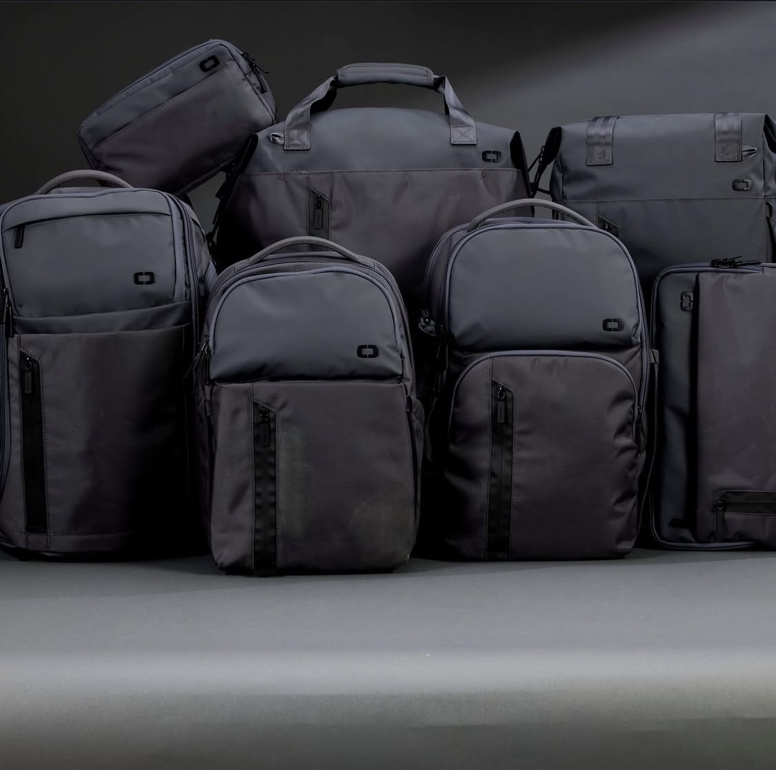 Eight black and gray bags of various sizes arranged on a surface, with a dark background.