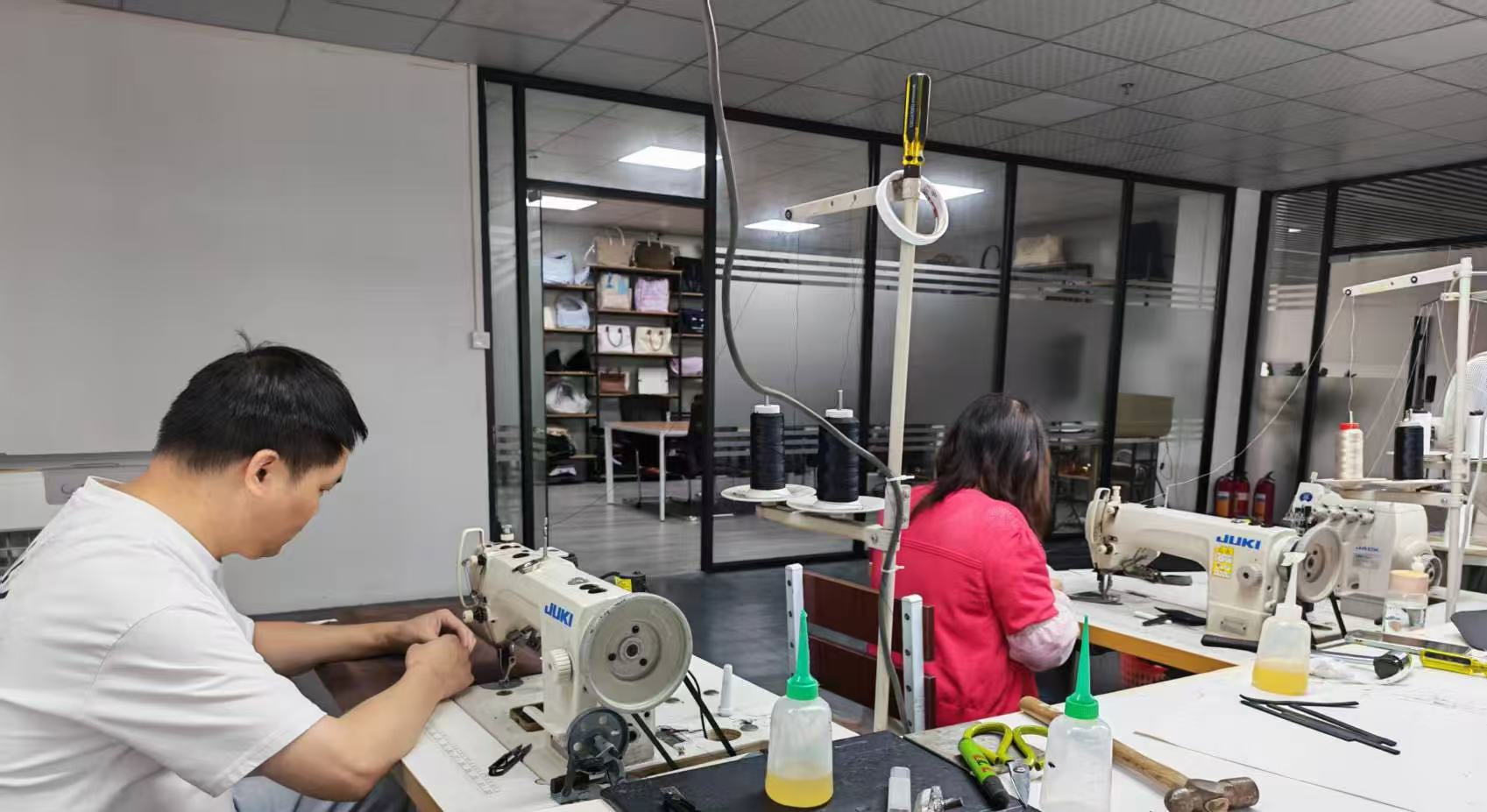 Two people working with sewing machines in a workshop, one in white and one in red, with equipment and supplies around them.