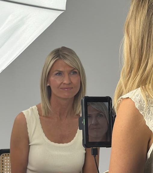 Woman with blonde hair looking at her reflection in a mirror, taking a selfie, with a gray wall background.