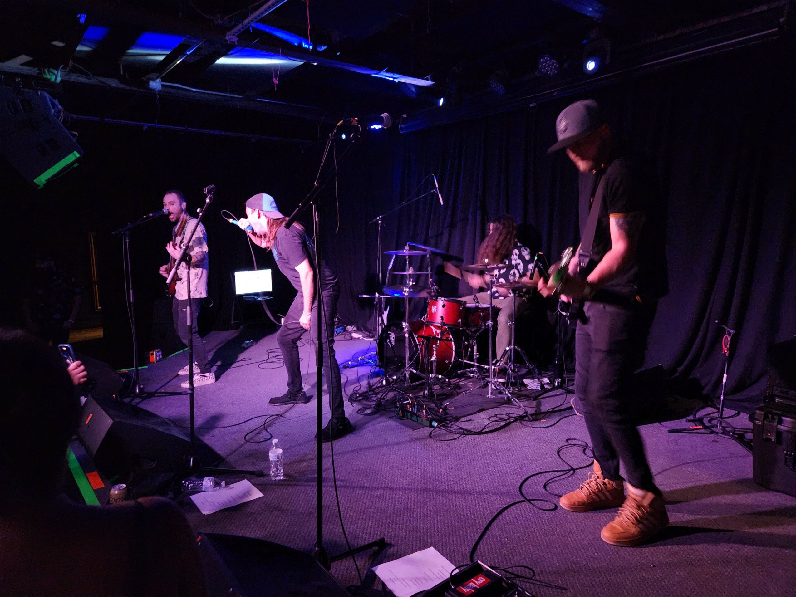 Band performing on stage with four members, one singing, others playing instruments, black curtain backdrop, stage lighting, music equipment.