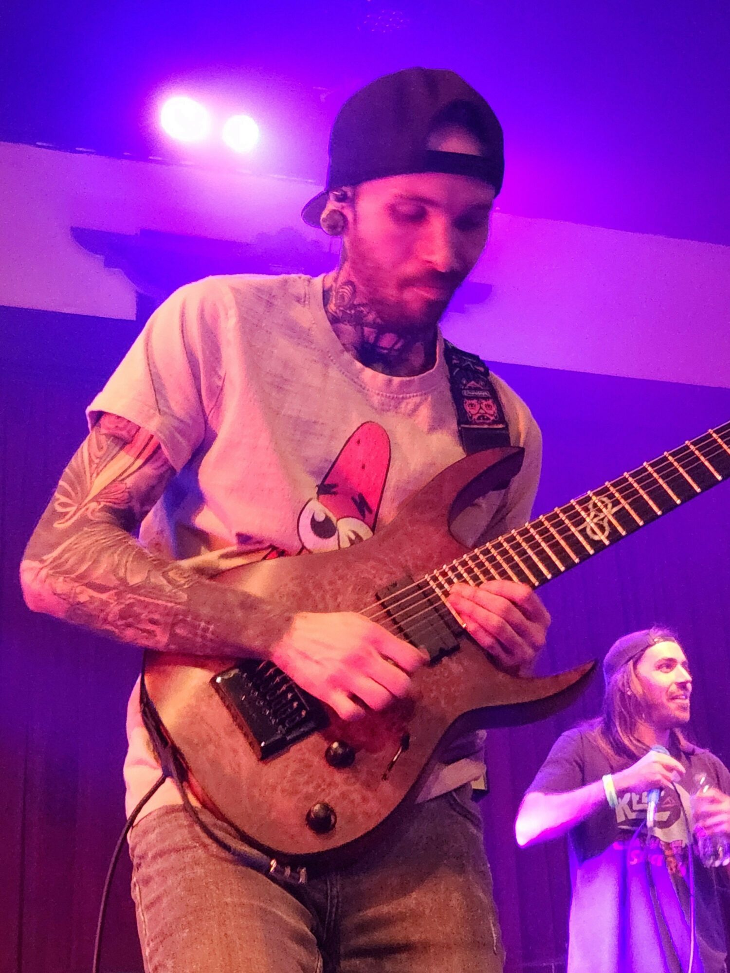 Person playing an electric guitar on stage with purple lighting, wearing a beanie and a cartoon character shirt.