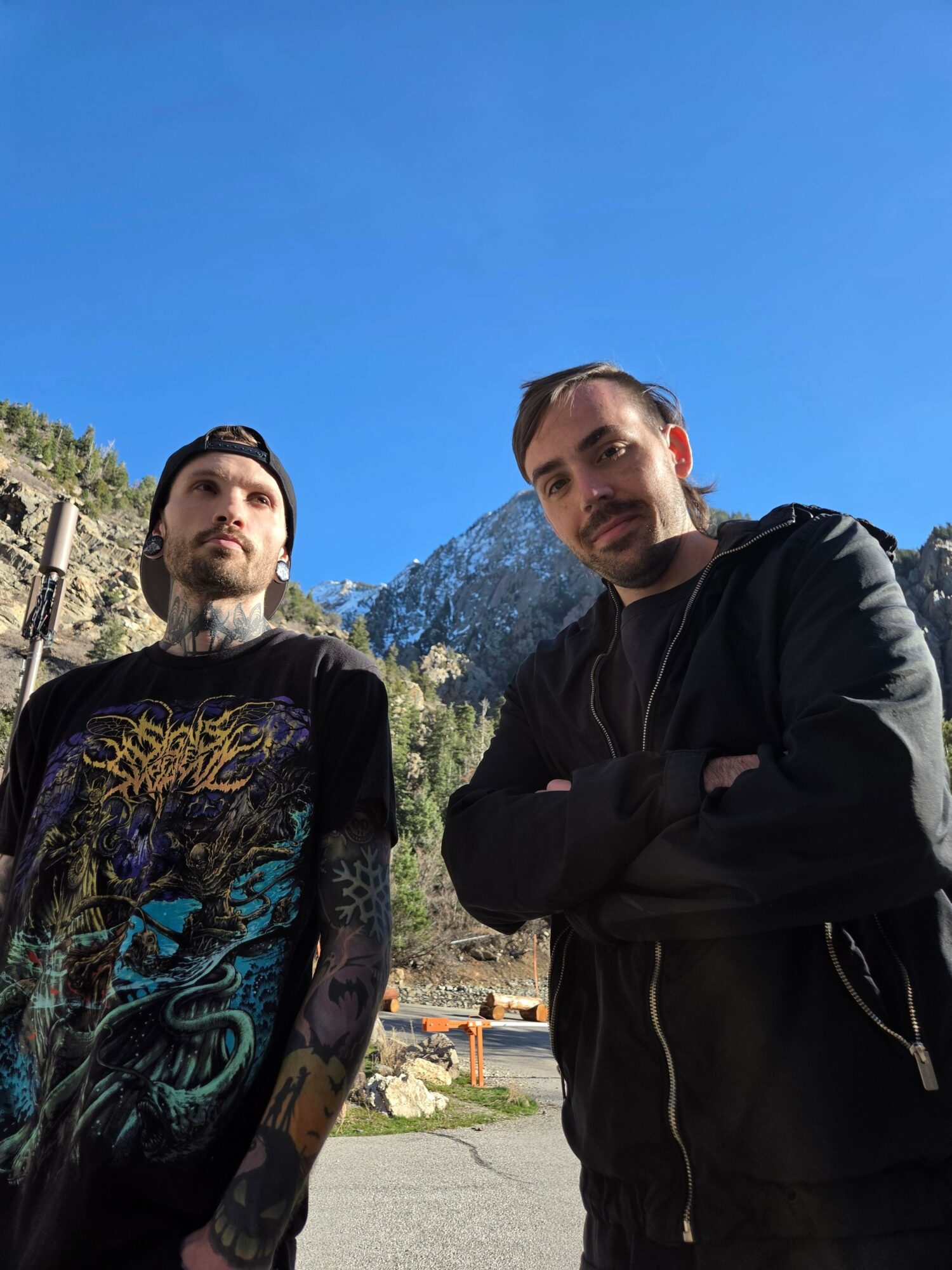 Two people standing outdoors with mountains and blue sky in background.