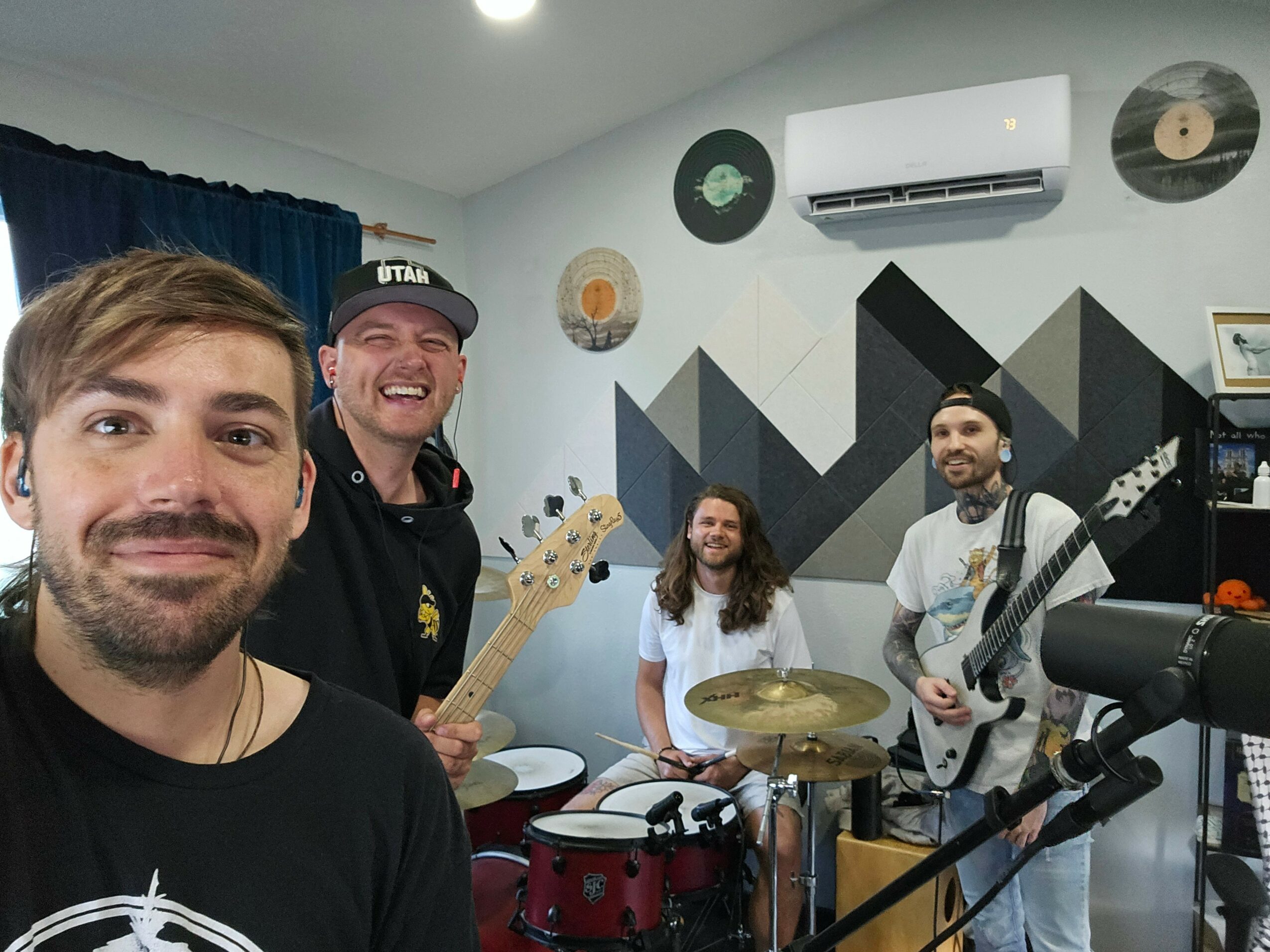 Four people in a music rehearsal room with instruments and wall decorations, smiling at the camera.