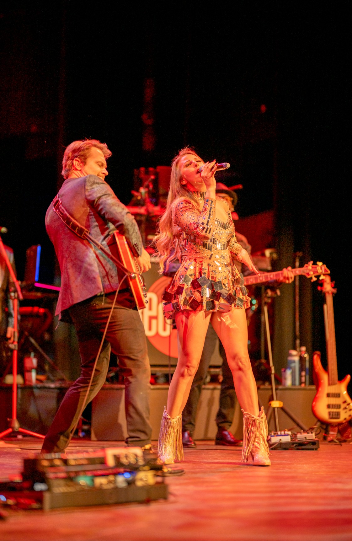 Two performers on stage, one playing guitar and the other singing, with musical instruments and equipment around them.
