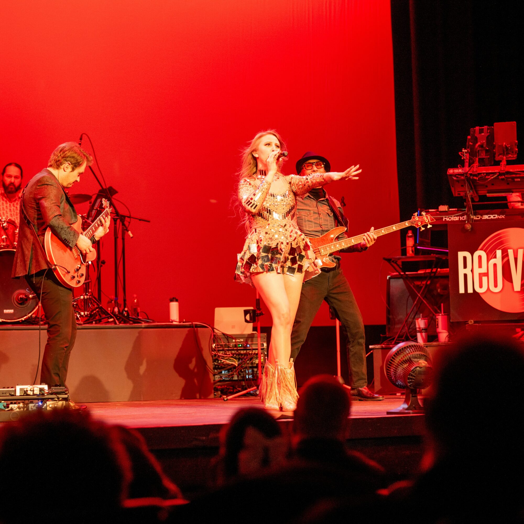 Musicians perform on stage with a red background, including a singer, guitarist, and drummer, with audience silhouettes in foreground.