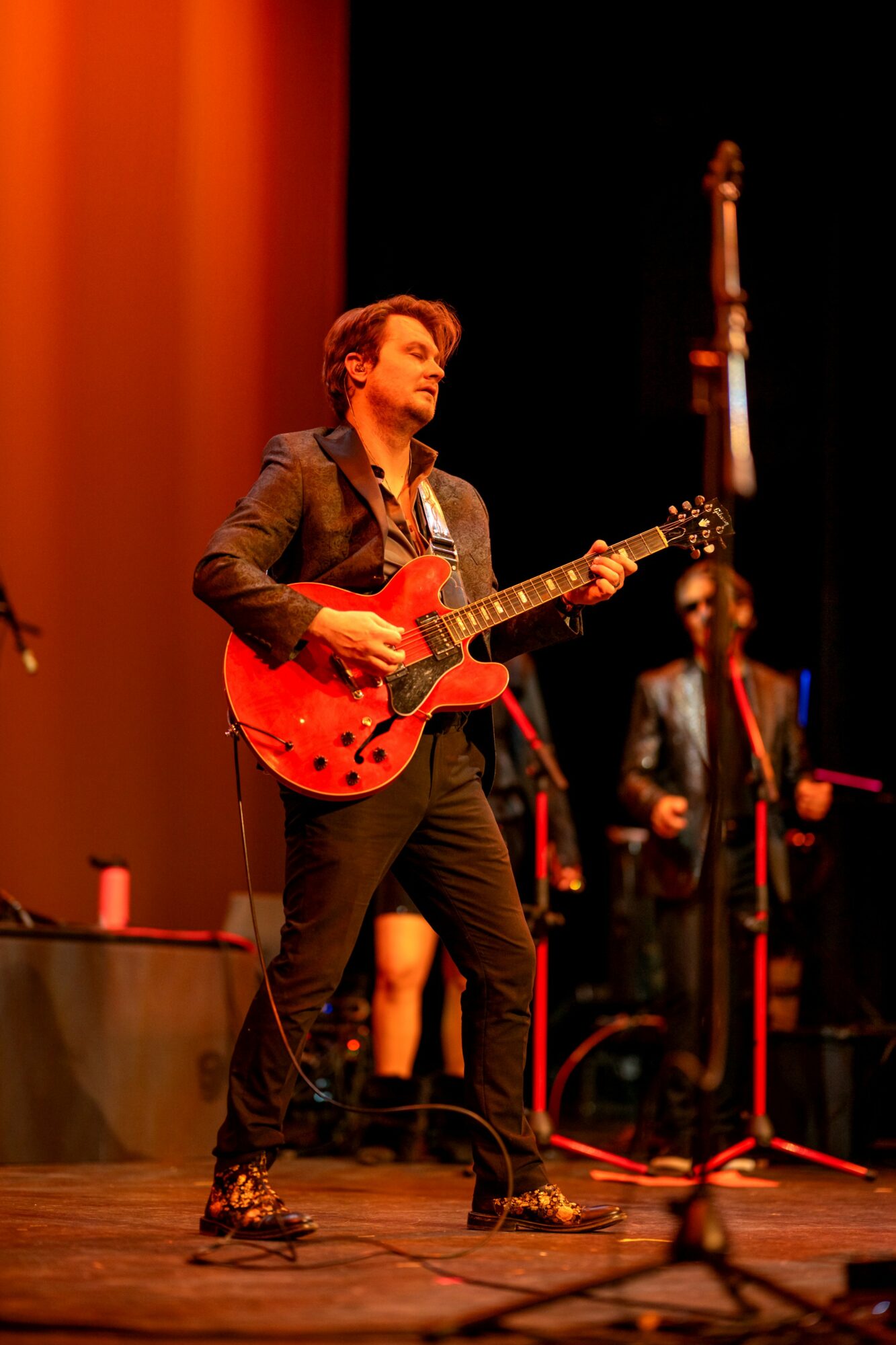 Musician playing red electric guitar on stage with band members in background, stage lighting, microphone in foreground.