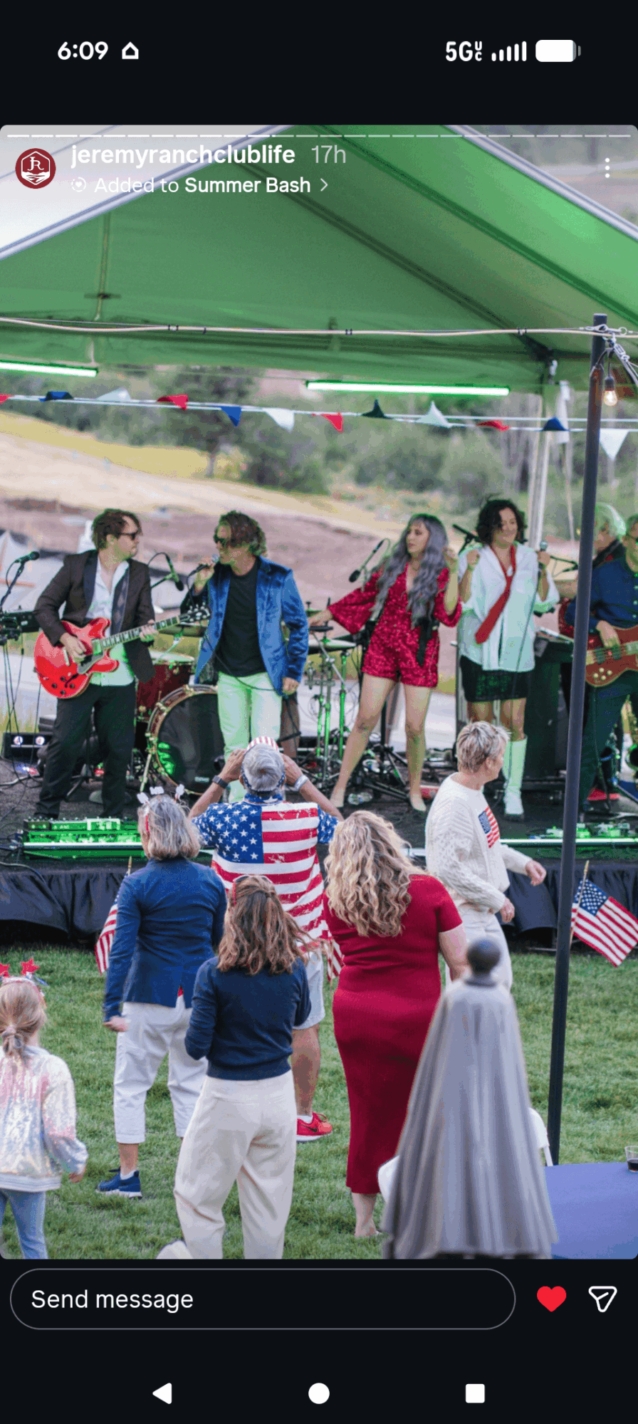 Group of children and adults gathered outdoors under a green tent, some holding American flags, at a summer event.