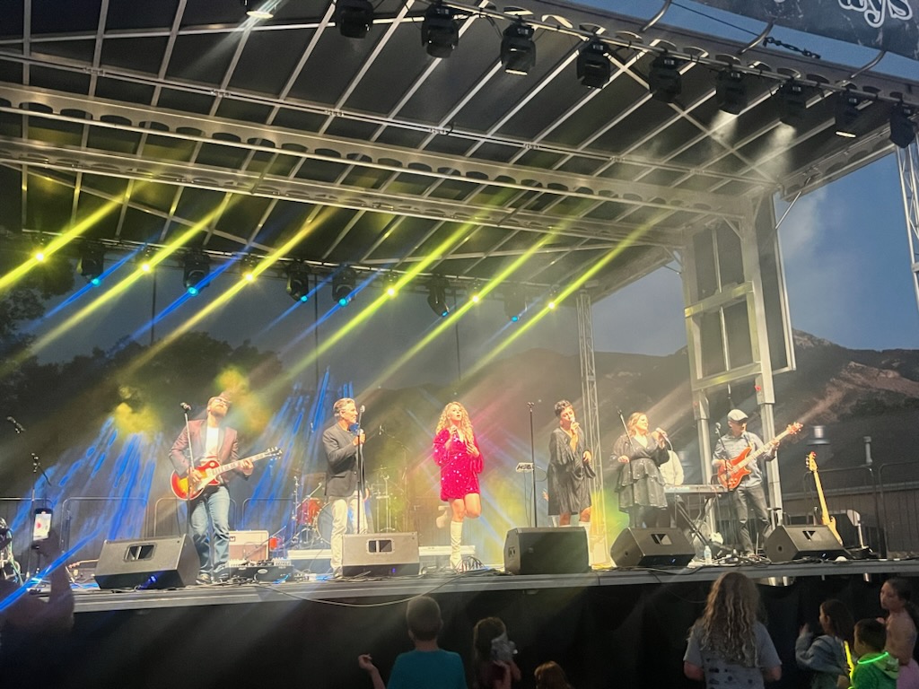 Band performs on stage with yellow and blue lighting, five musicians, some with guitars, outdoors during daytime.