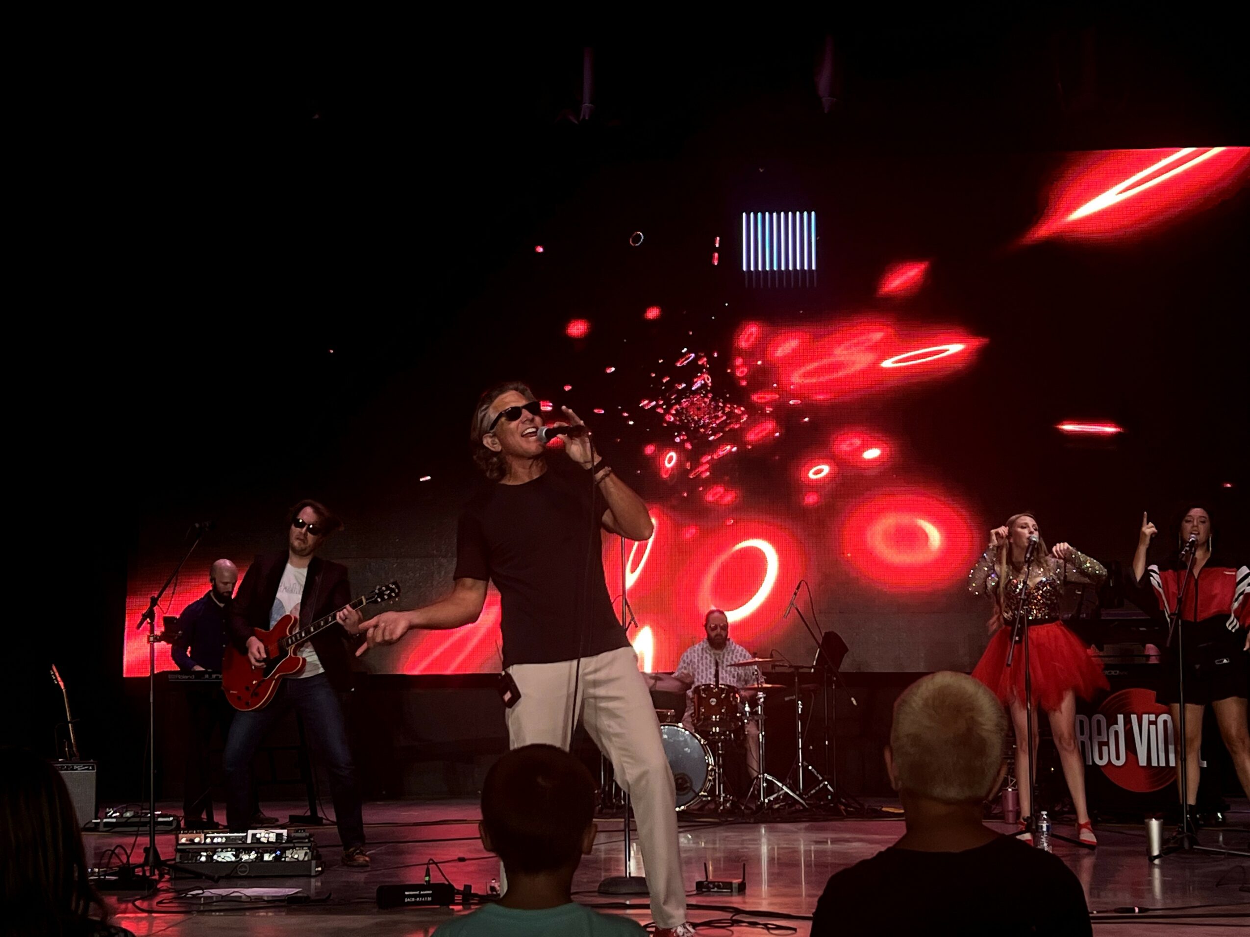 Band performs on stage with red and black background, singer in center holding microphone, musicians playing instruments.